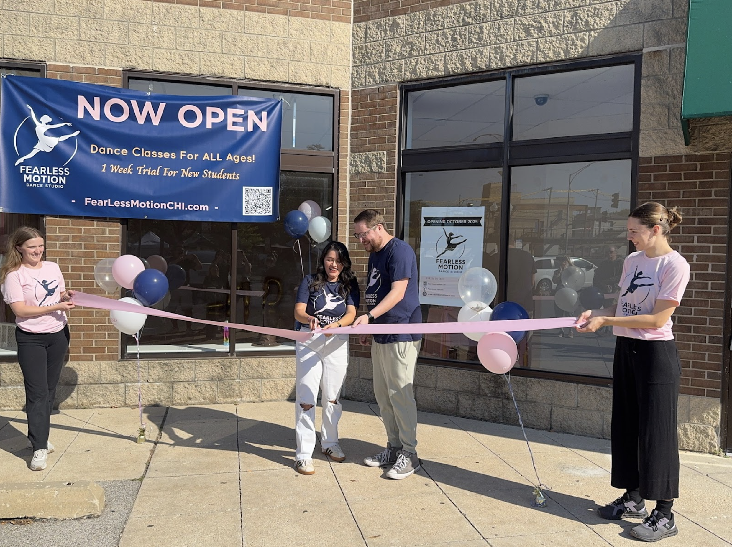 People celebrating a grand opening outside a dance studio called Fearless Motion, with a ribbon-cutting ceremony, pink, blue, white, and purple balloons, and a large banner that reads 'NOW OPEN' with information about dance classes.