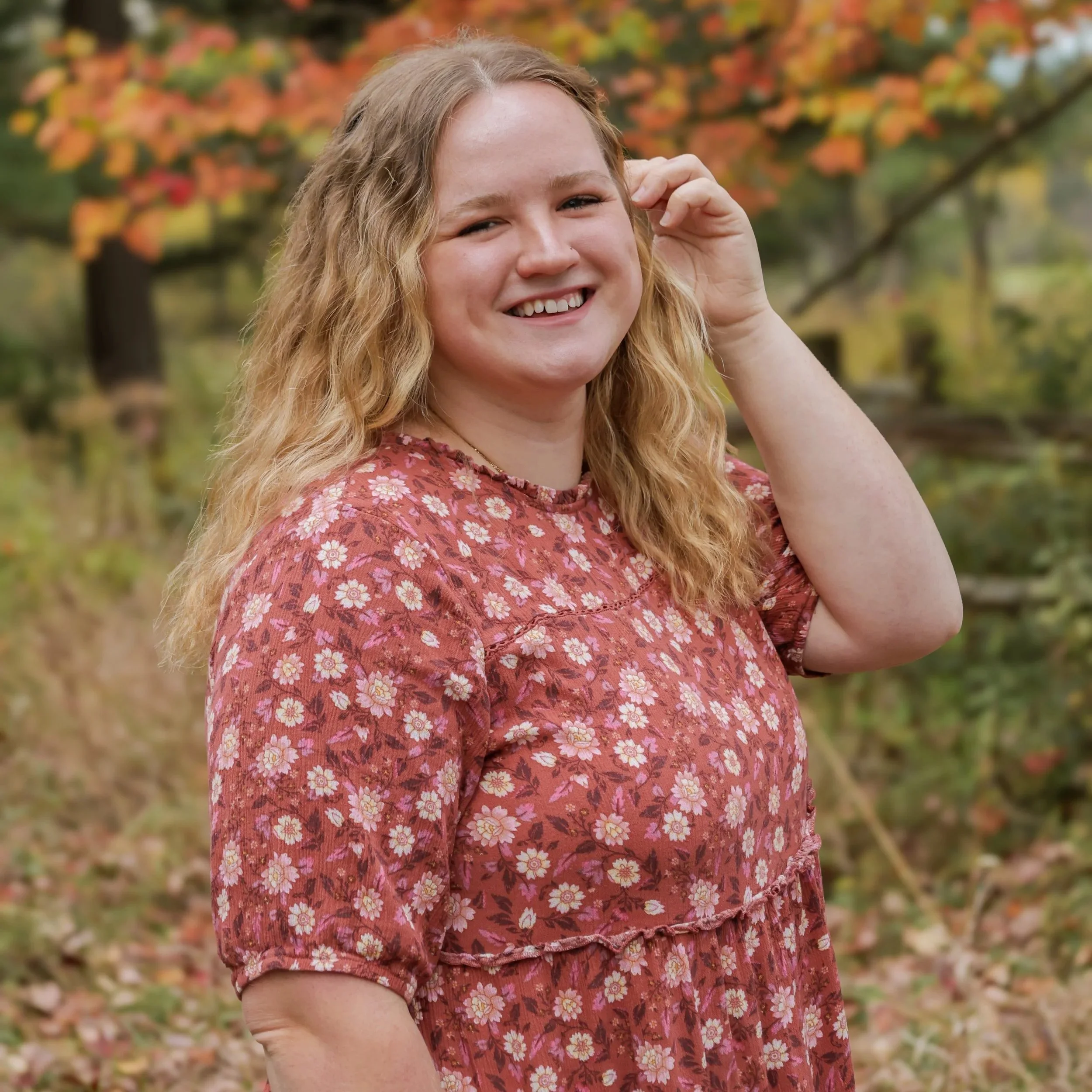 A smiling young woman with long, curly blonde hair wearing a reddish floral dress standing outdoors with autumn foliage in the background.