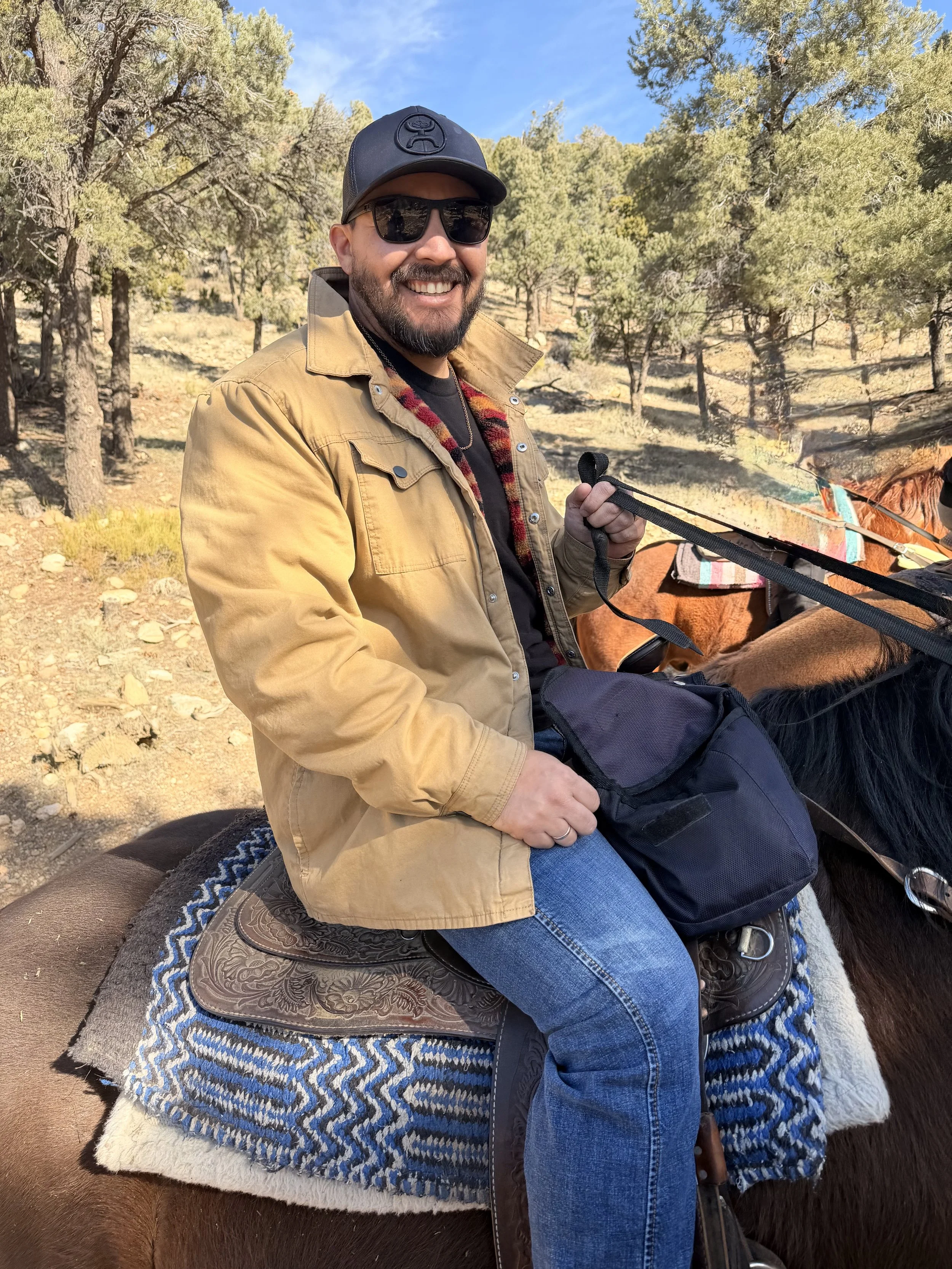 Man riding a horse outdoors in a wooded area, smiling, wearing sunglasses, a black cap, a tan jacket, and blue jeans, holding the reins and a black bag.