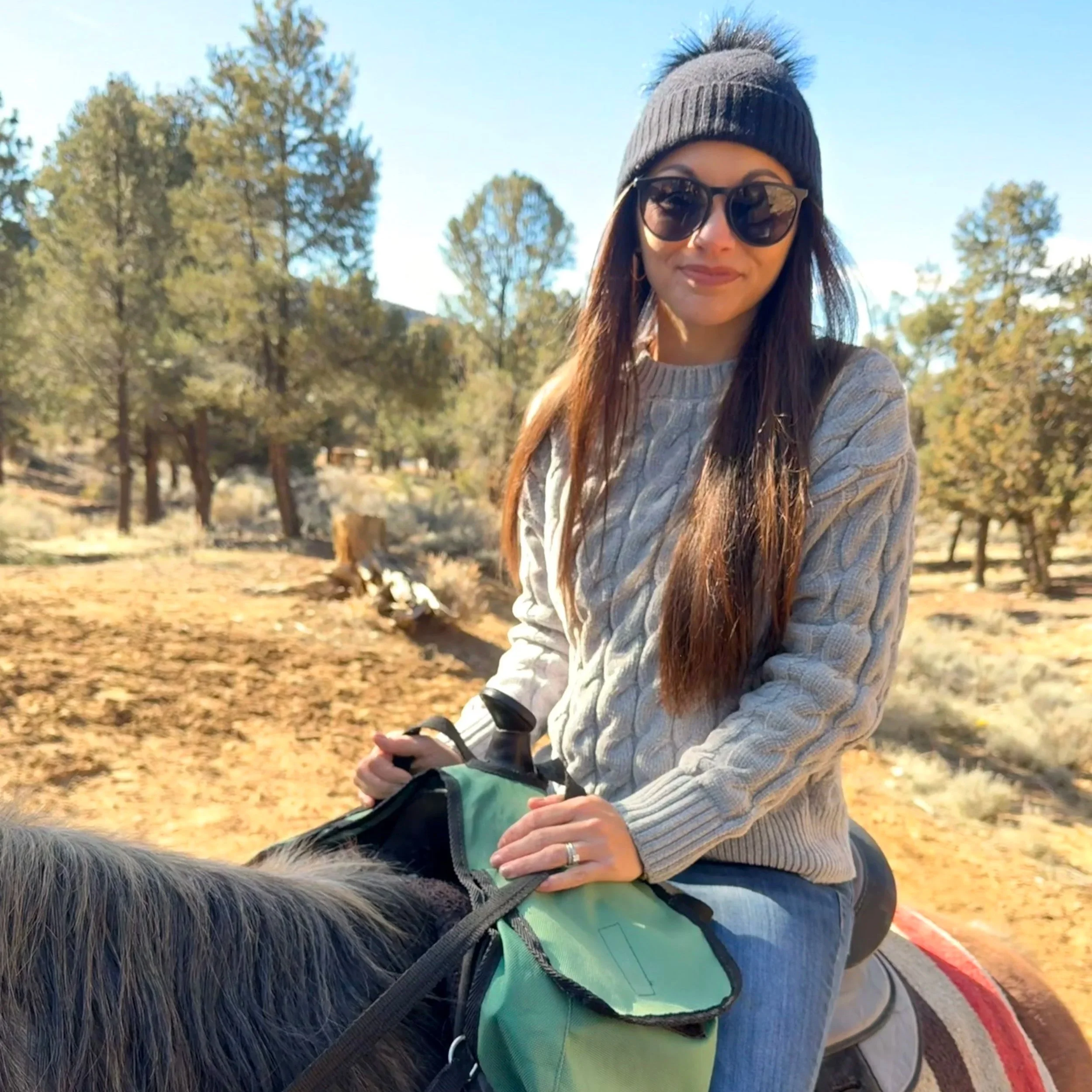 A woman with long brown hair wearing sunglasses, a black beanie, and a beige sweater riding a horse on a trail surrounded by trees on a sunny day.