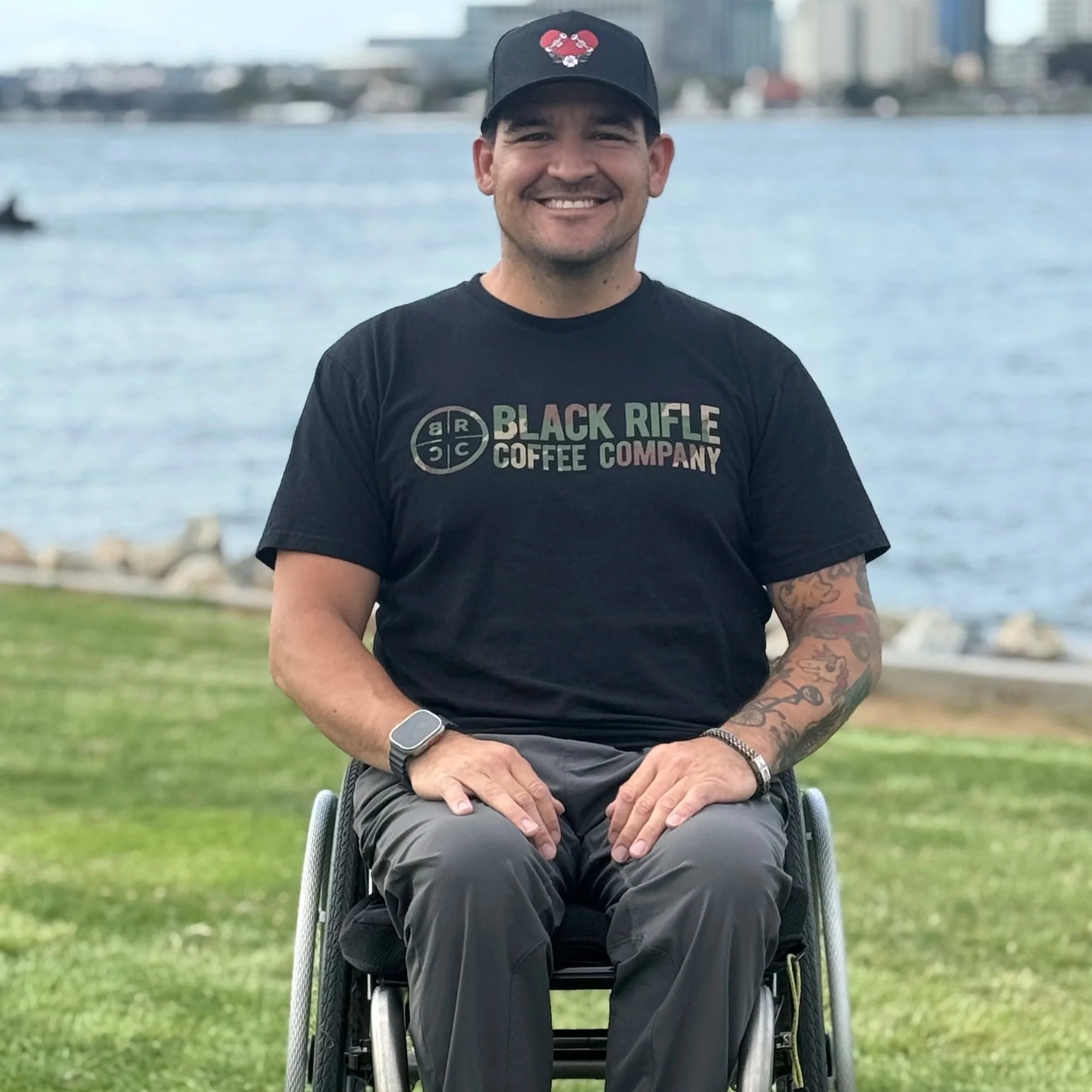 A smiling man in a wheelchair outdoors near a body of water, wearing a black t-shirt and baseball cap.