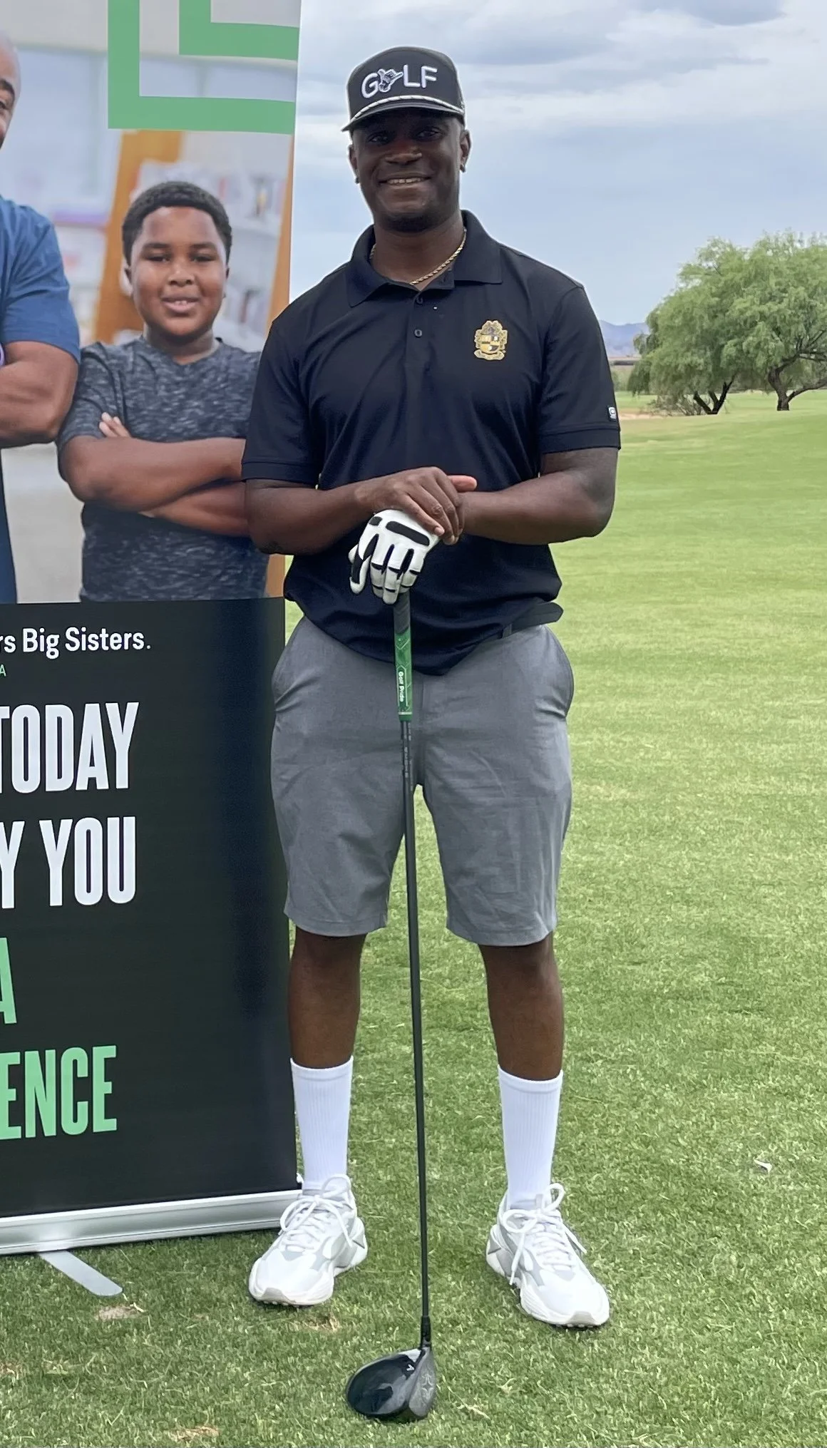 A man standing on a golf course holding a golf club, wearing a black polo shirt, gray shorts, white sneakers, and white socks, with a golf glove on his left hand. There are trees in the background and a cloudy sky.