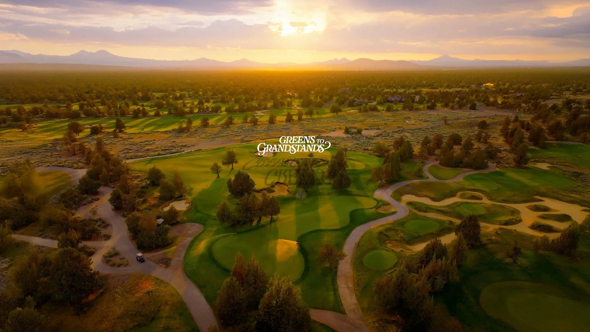 Aerial view of a scenic golf course at sunset with winding paths, trees, sand traps, and a logo for 'Greens to Grandstands' in the center.