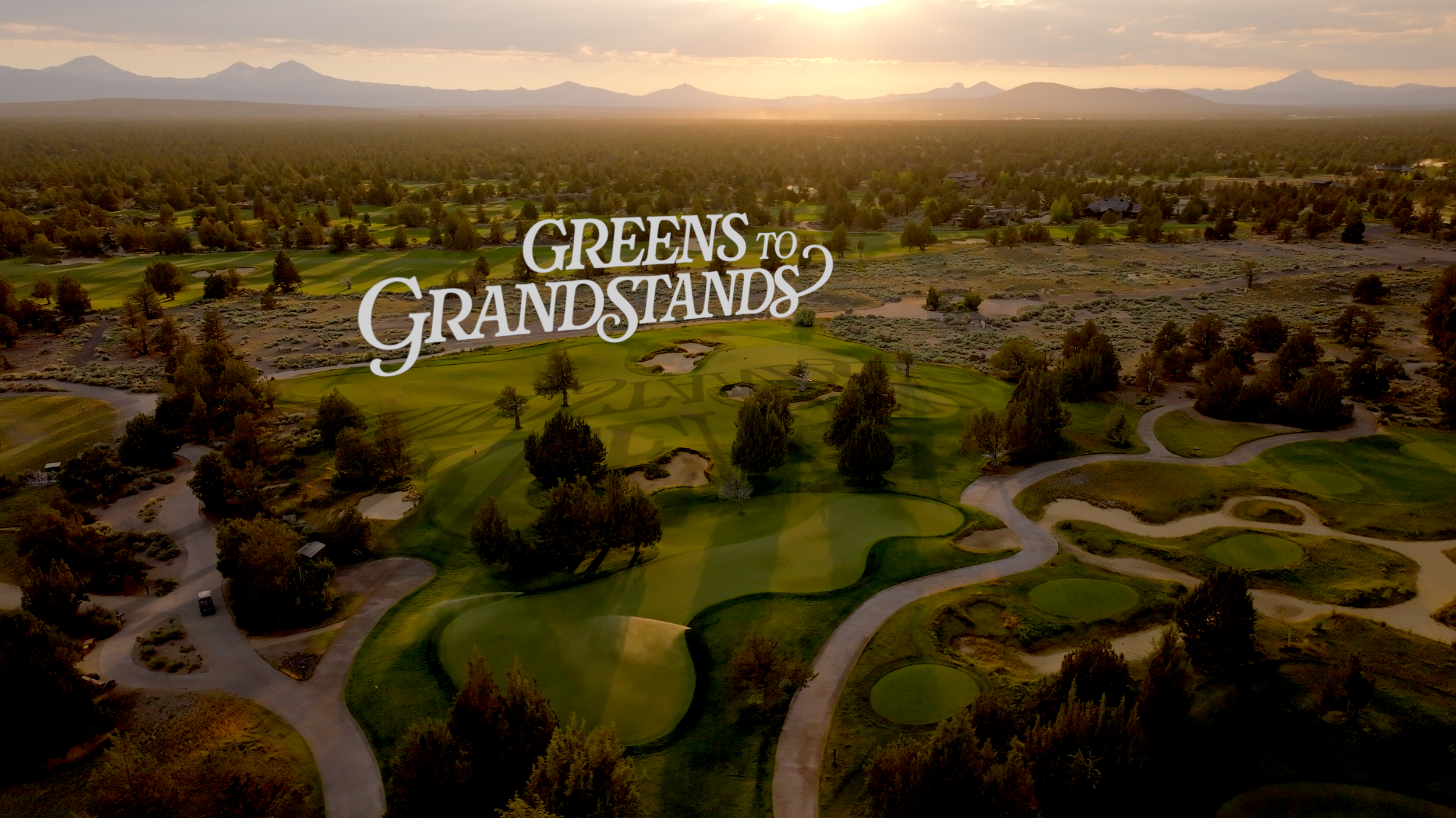 Aerial view of a golf course surrounded by trees, with mountains in the background at sunset. Text overlay says 'Greens to Grandstands'.