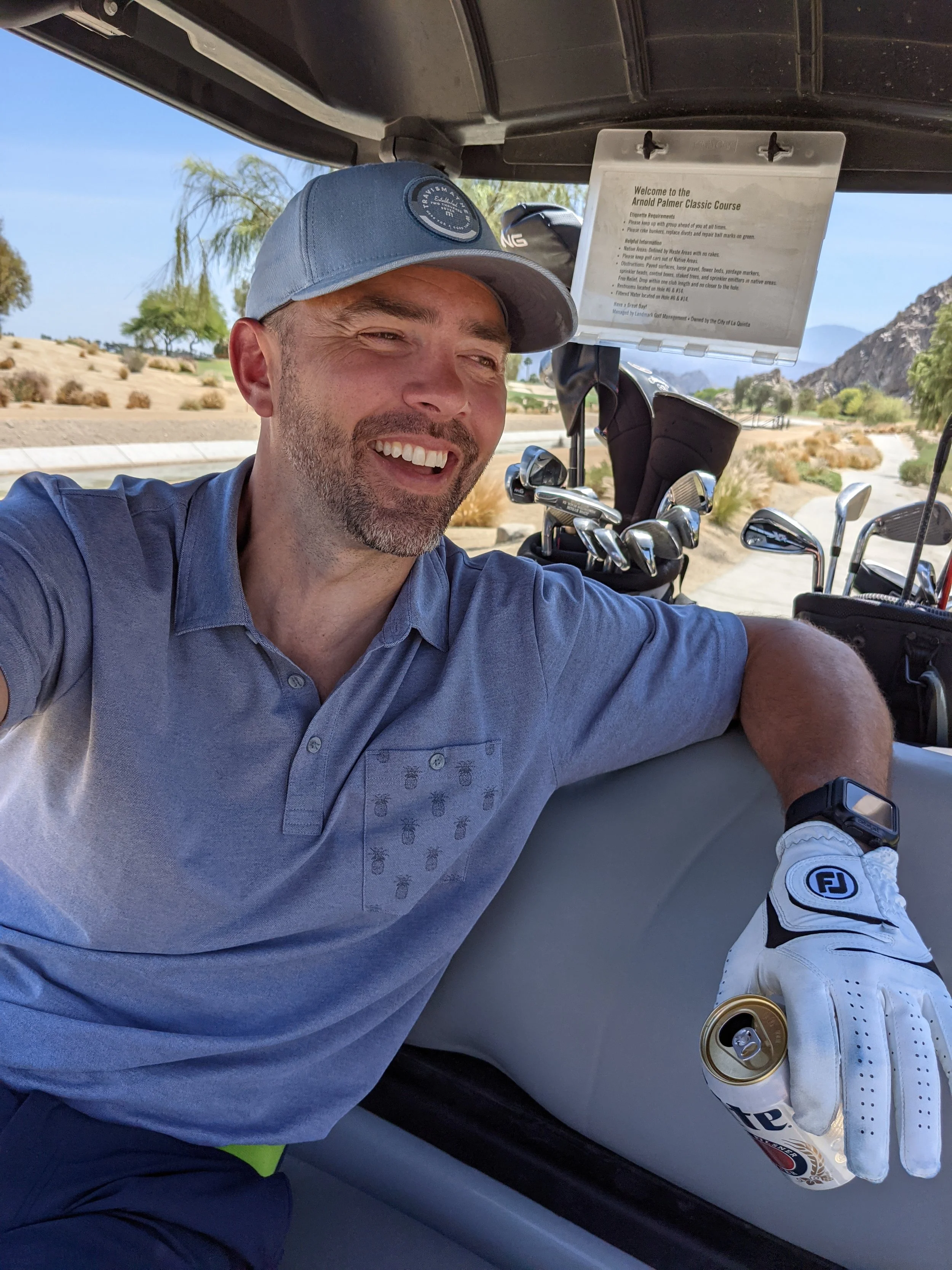 A man in golf attire taking a selfie inside a golf cart at a golf course with trees, mountains, and a clear sky in the background.