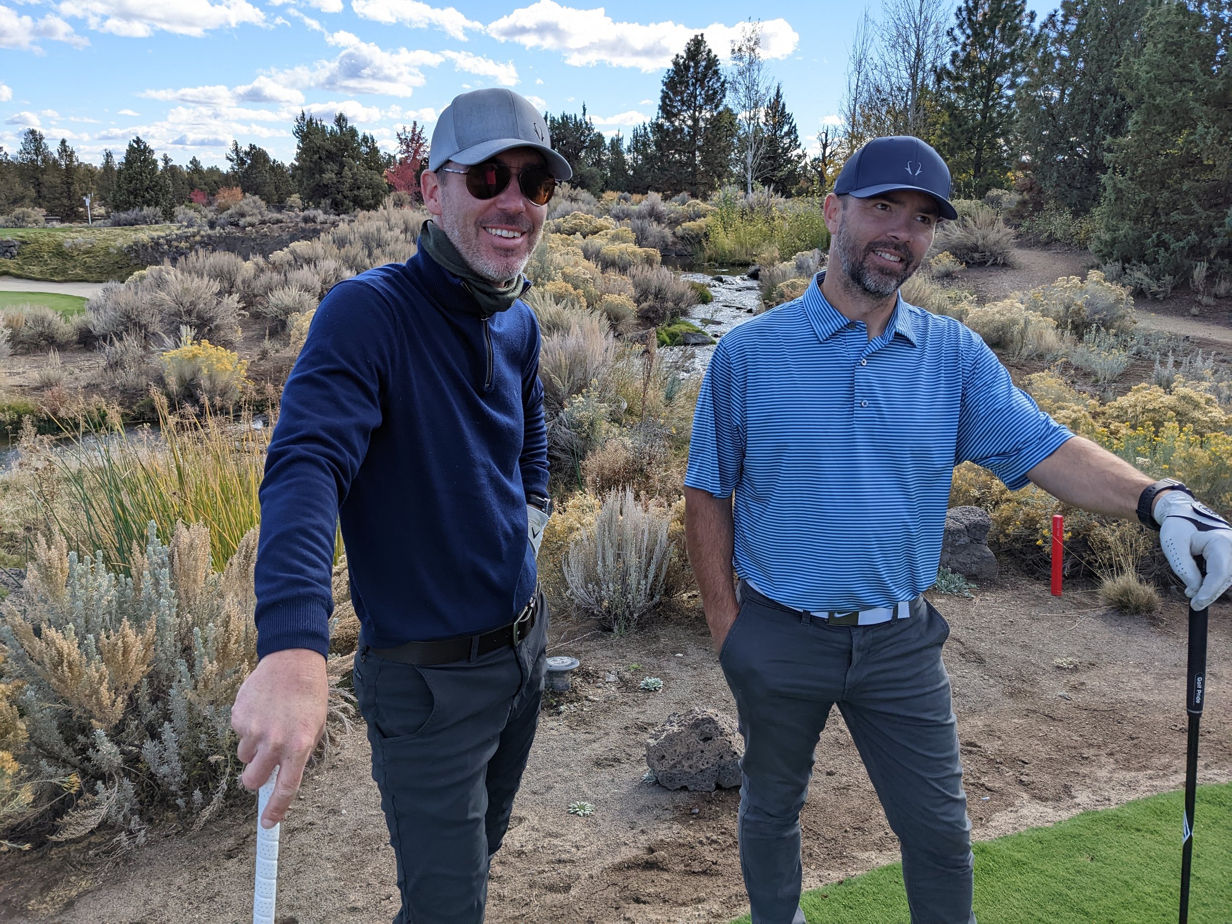Two men standing on a golf course surrounded by shrubs, trees, and a small creek, with blue skies overhead.