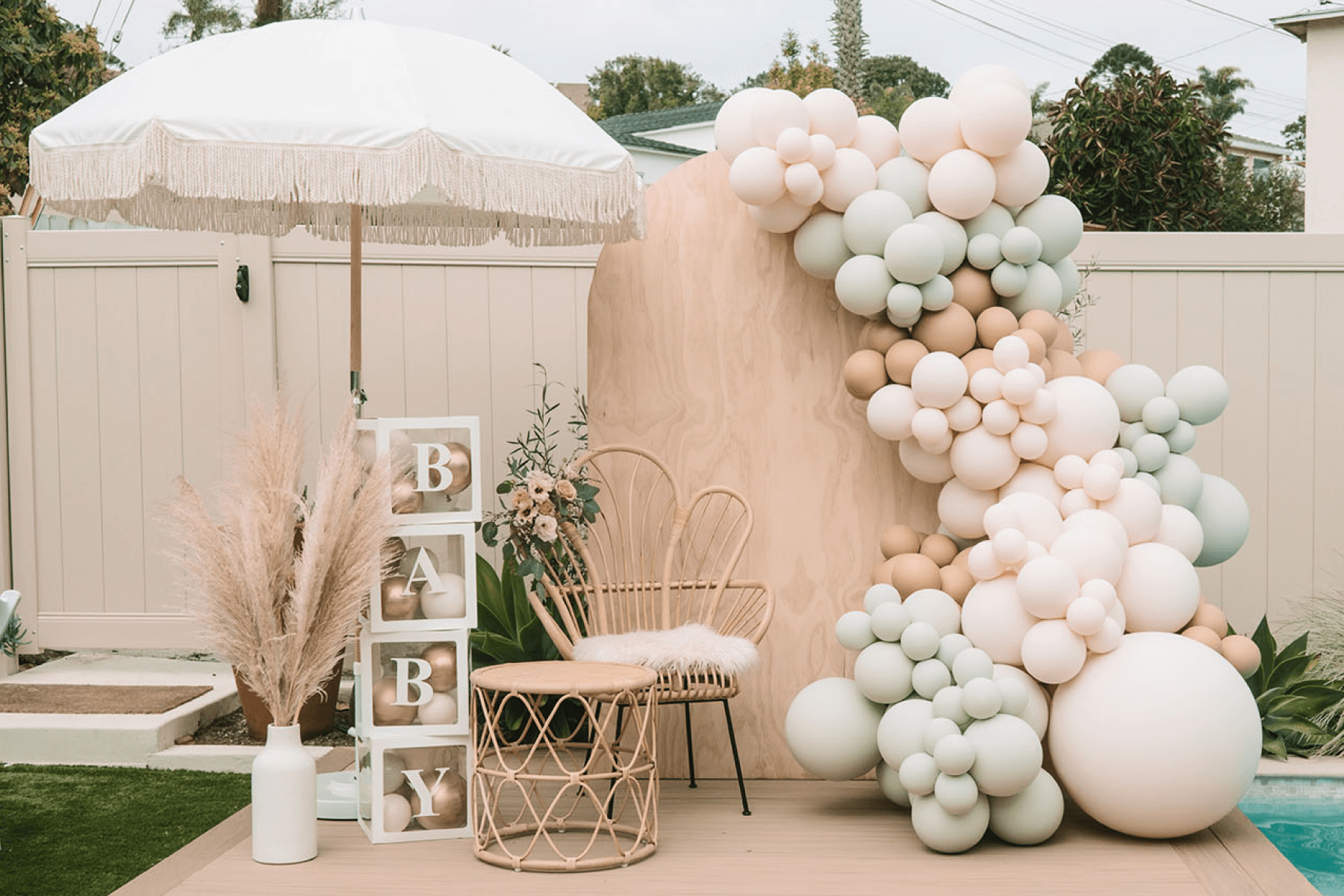 Neutral outdoor baby shower backdrop with ivory and sage balloons, woven chairs, and pampas grass.
