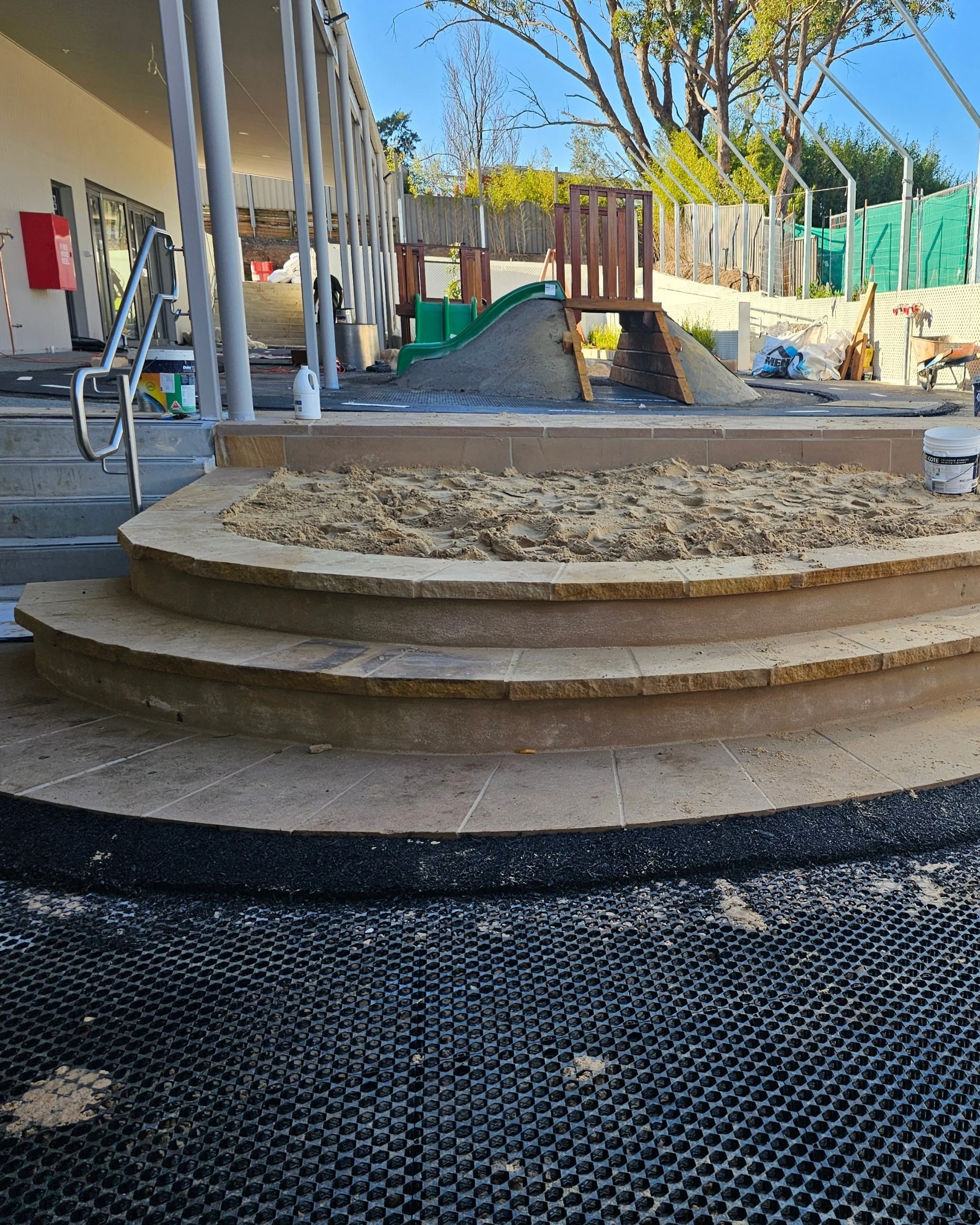 Under construction outdoor playground area with sand, steps, and a slide with a green top, wooden structure, and dirt mounds.