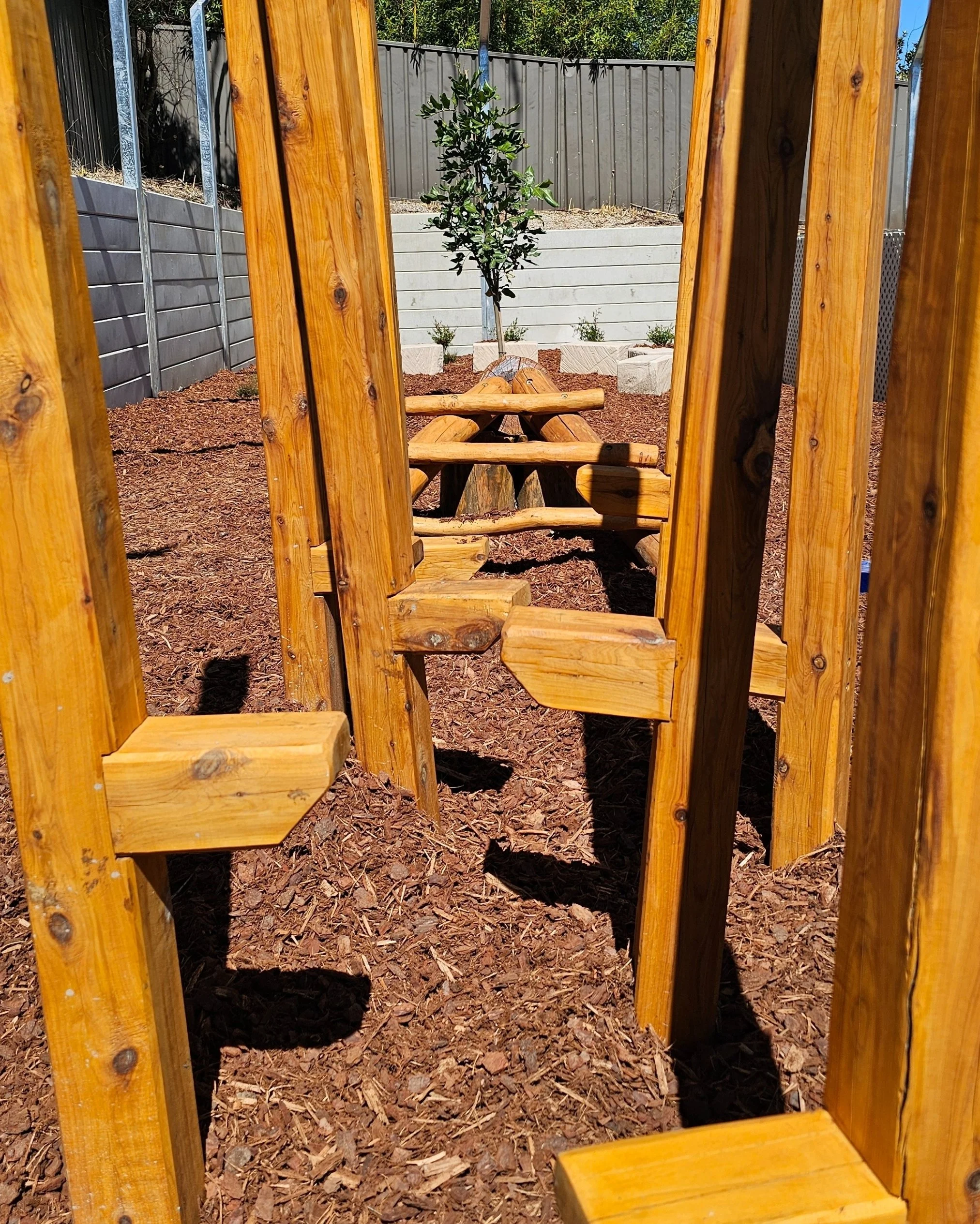 A wooden playground structure with a tunnel and seating area, view through vertical wooden bars.