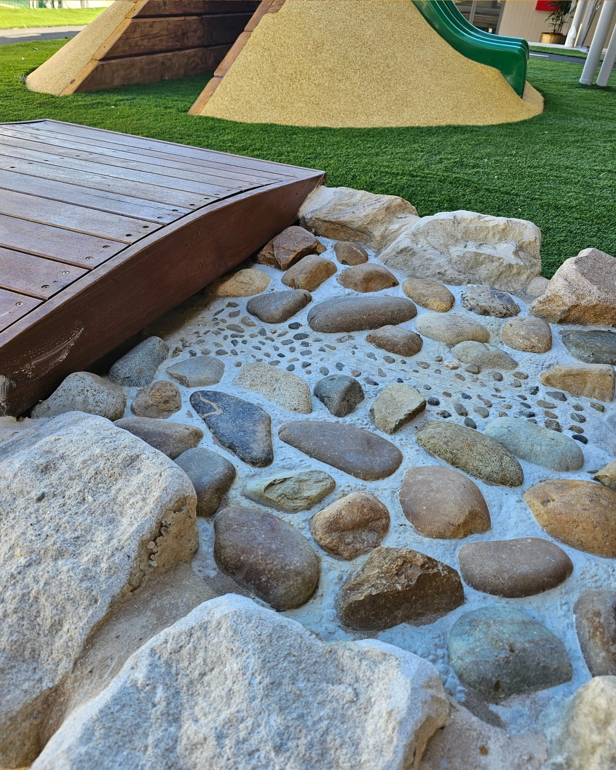Close-up of a small decorative rock garden with various stones and pebbles around a wooden bridge, with artificial grass and playground equipment in the background.