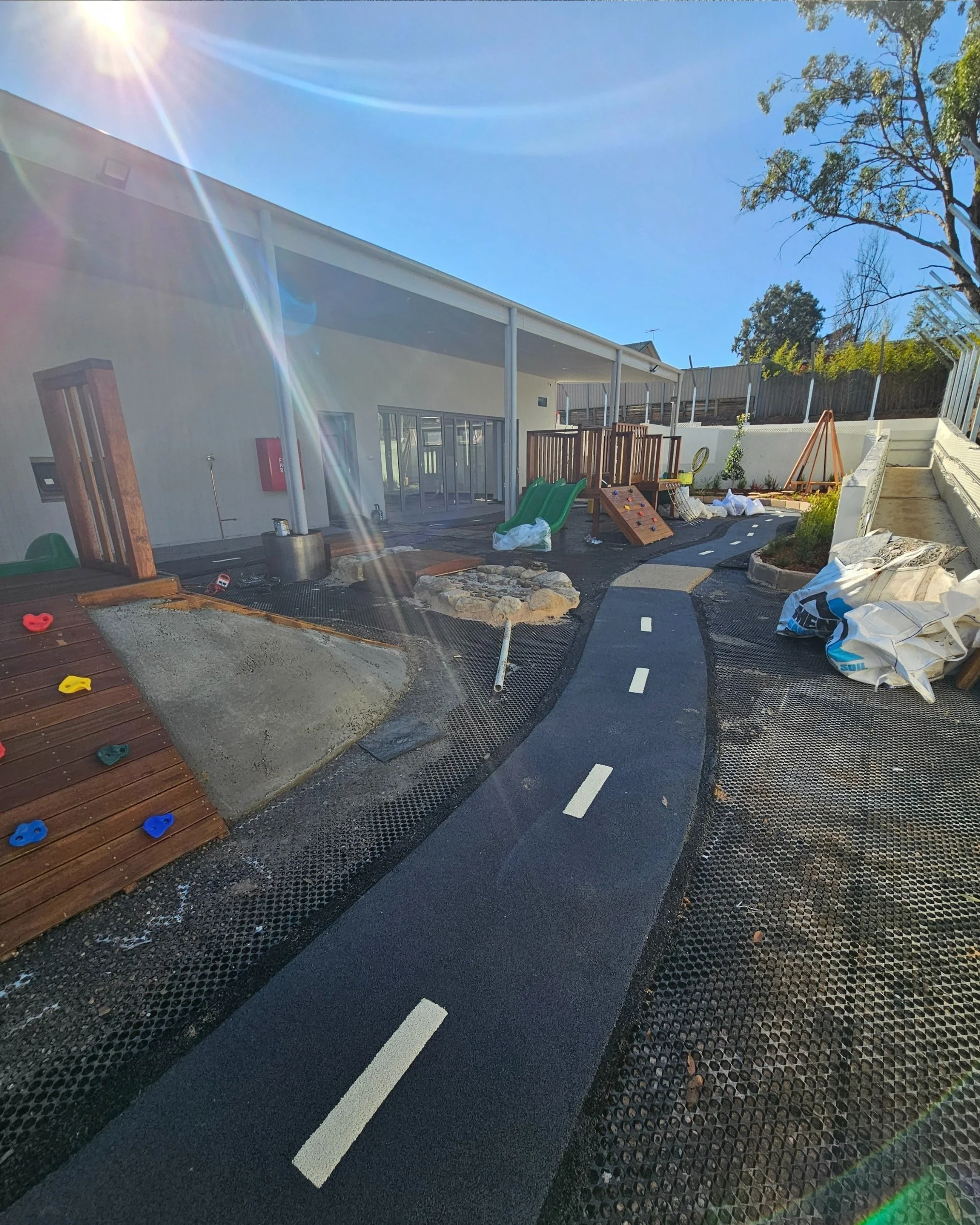Under construction backyard with a small asphalt walking path, wooden play structures, rocks, and construction materials under a bright sun in a sunny day.