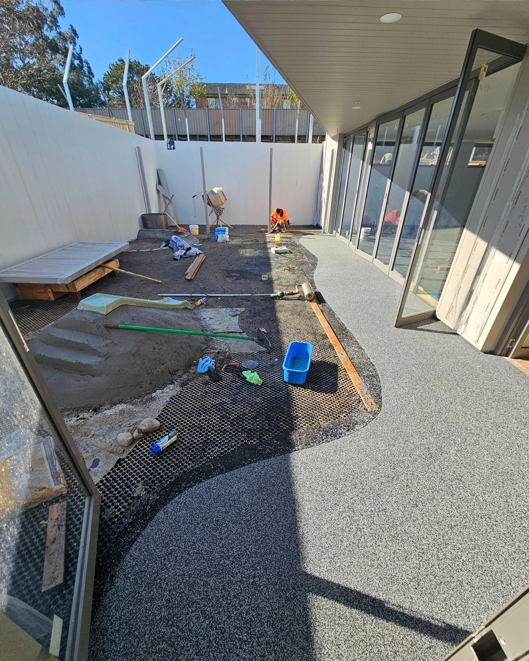 Construction work on a backyard patio with workers, tools, and materials. Partially completed flooring with some areas still being worked on.