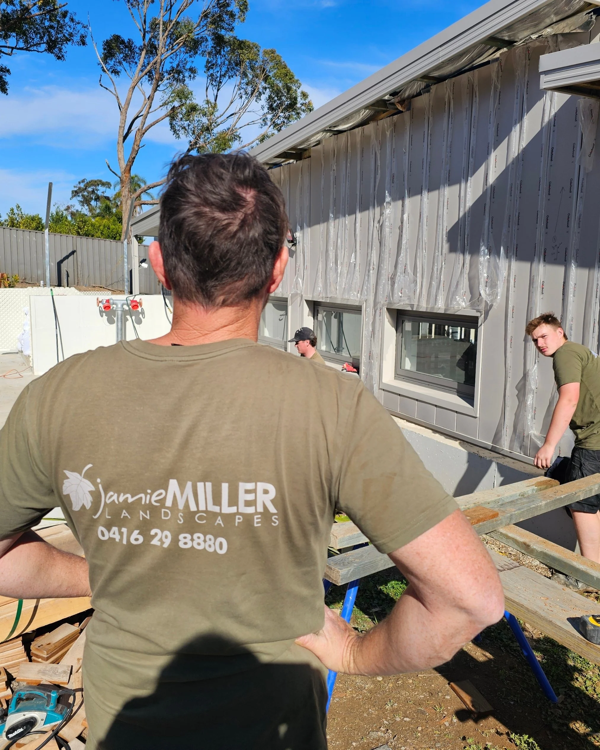 A man with his back to the camera wearing a beige T-shirt with 'Jamie Miller Landscapes' and a phone number printed on it, standing outdoors at a construction site. There are two young men working on a building under construction in the background, w