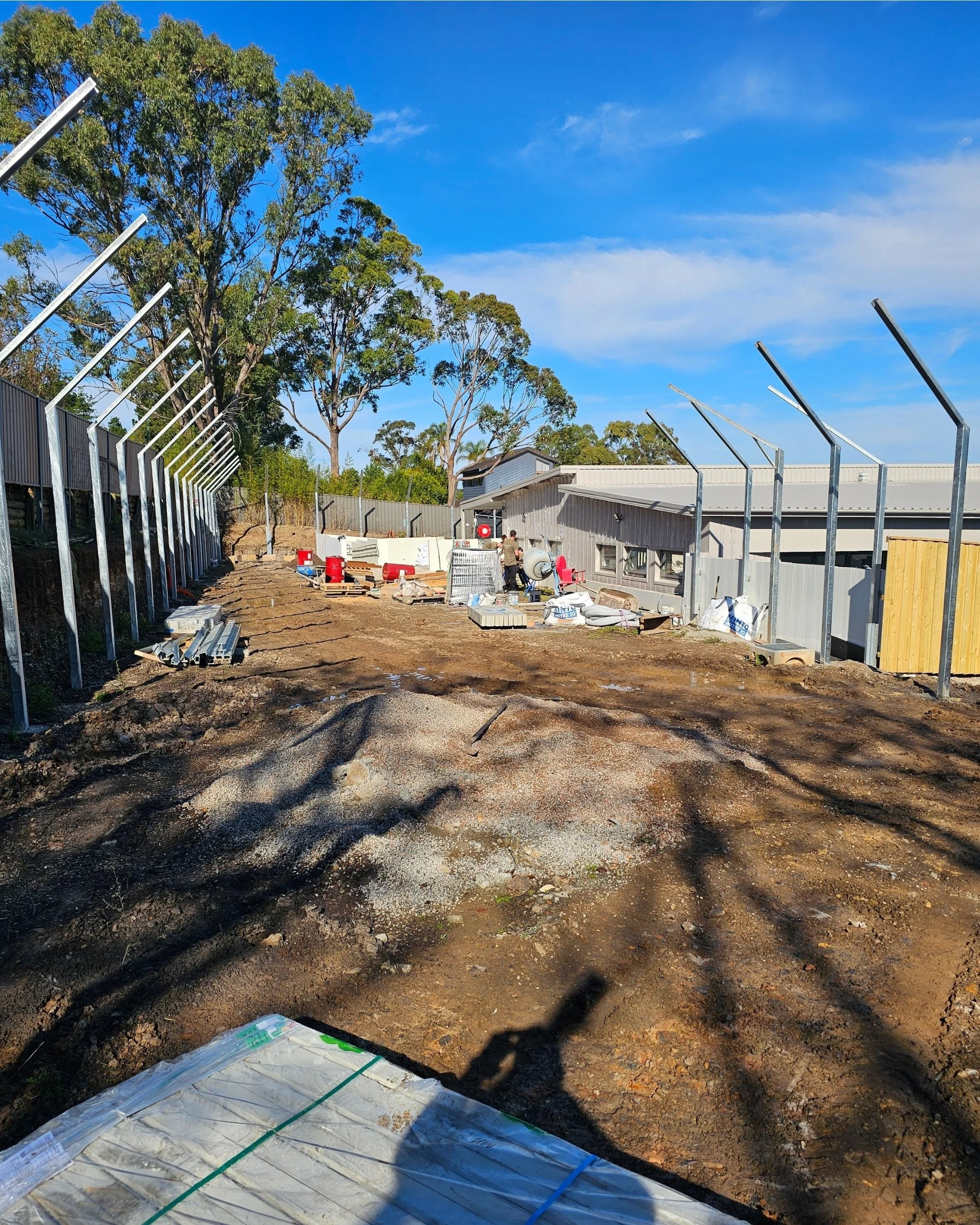 Construction site with metal fence posts, building materials, and equipment under a clear blue sky with trees in the background.