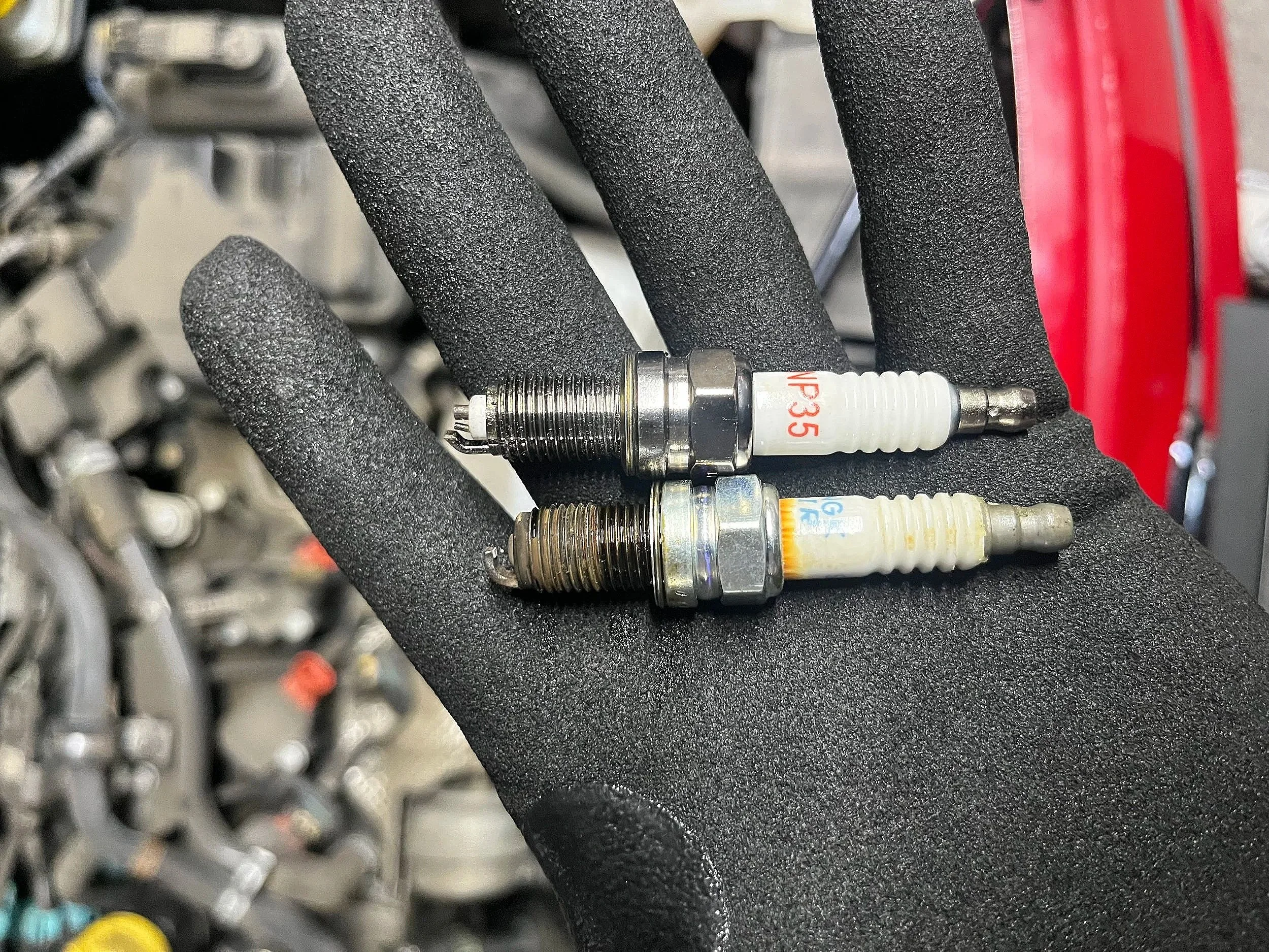 Two spark plugs, one new and one used, held on a black glove in front of a blurred background of a car engine.