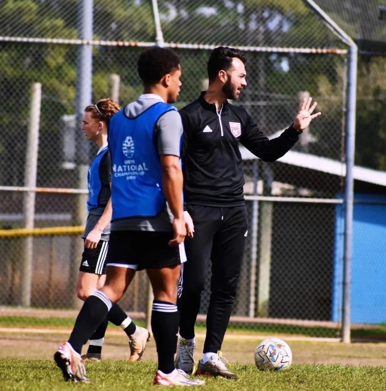 A soccer coach giving instructions to players on the field during practice or game, with a soccer ball on the grass and a chain-link fence in the background.