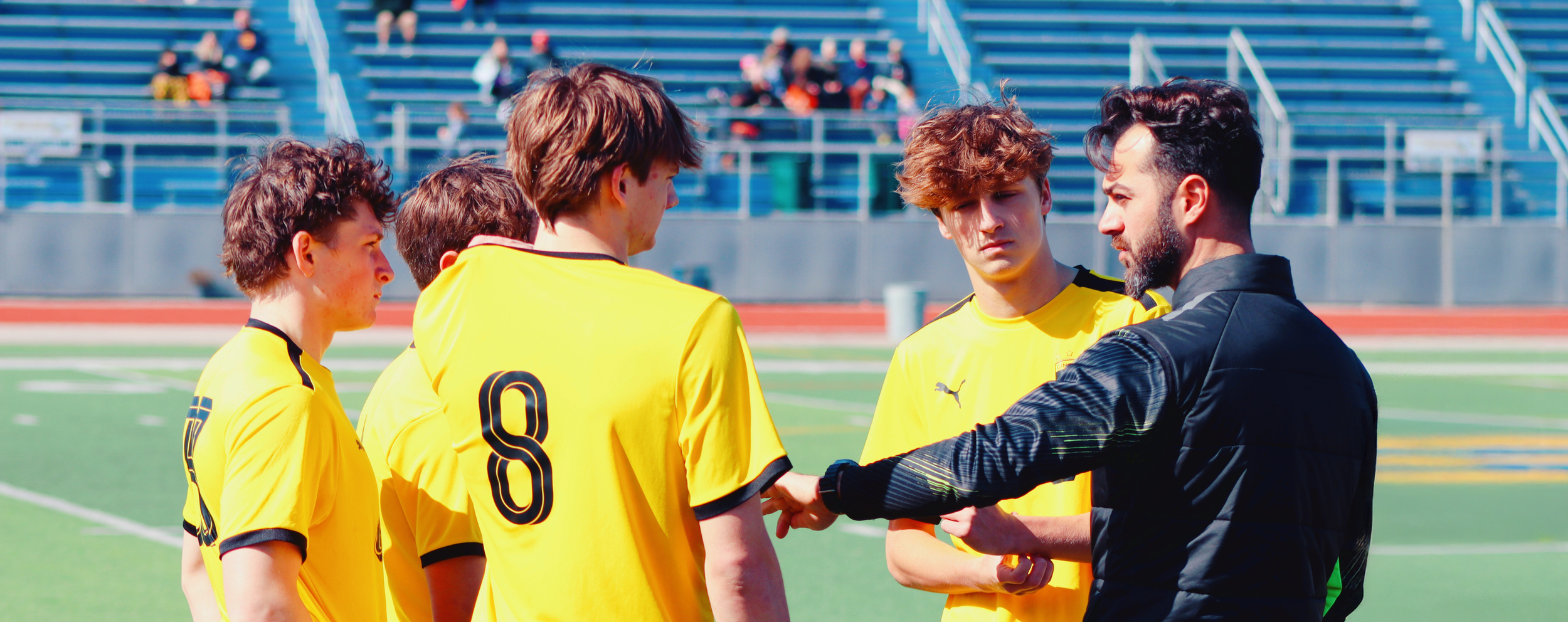 A group of young male soccer players in yellow jerseys listening to their coach on a soccer field.