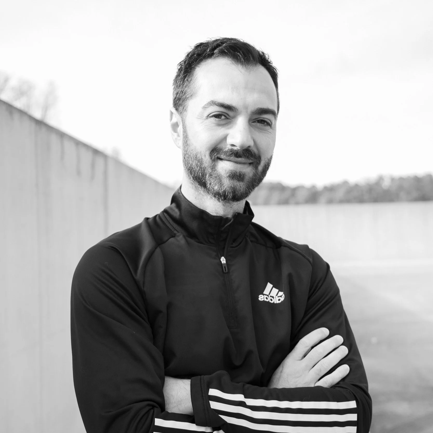Man in Adidas jacket standing outdoors with arms crossed, monochrome photo.