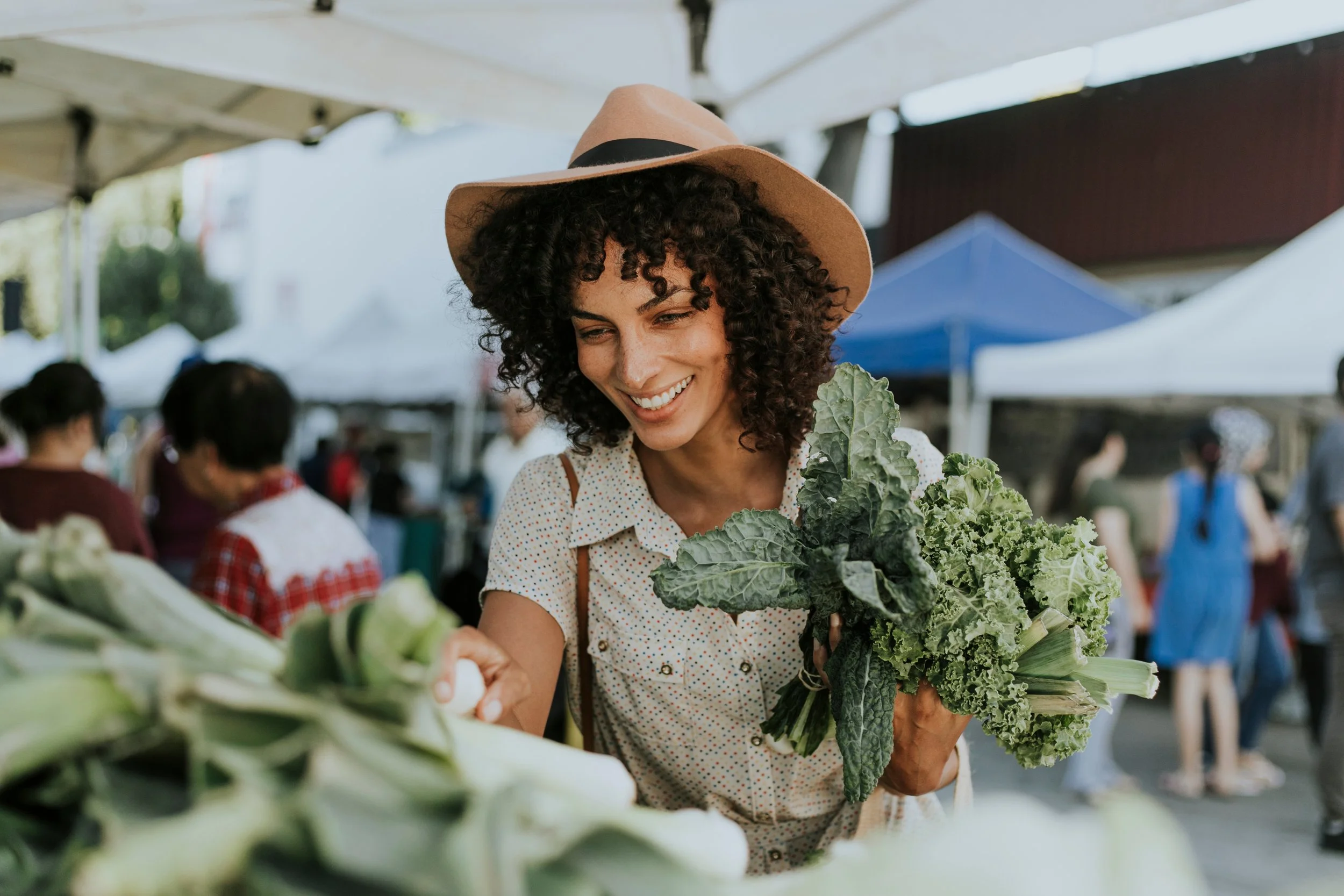 Woman shopping at a farmer's market, holding kale and wearing a brown hat.