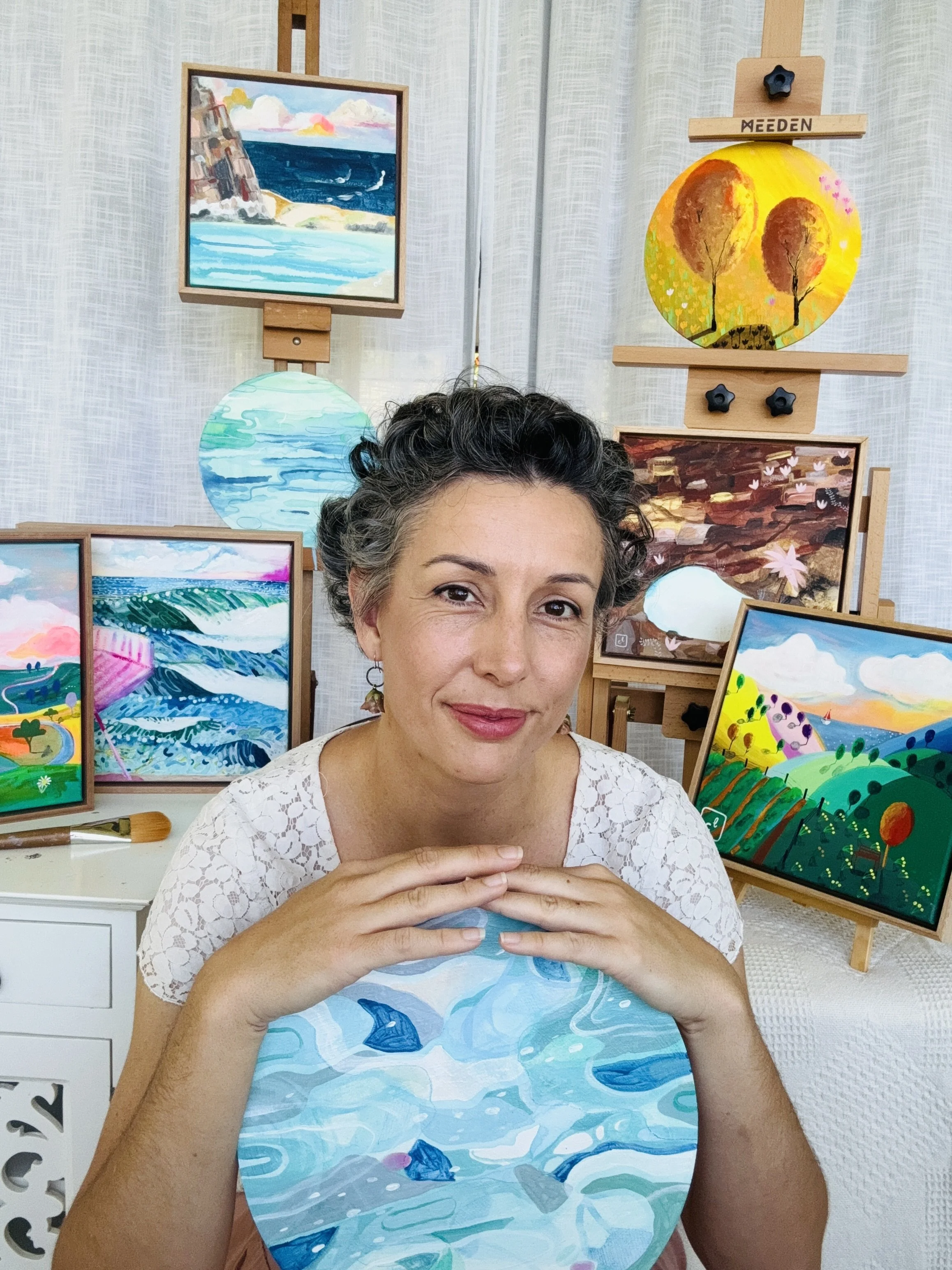 A woman with short curly hair in a white lace top, sitting in front of a display of colorful landscape paintings, holding a painting of an abstract water scene.