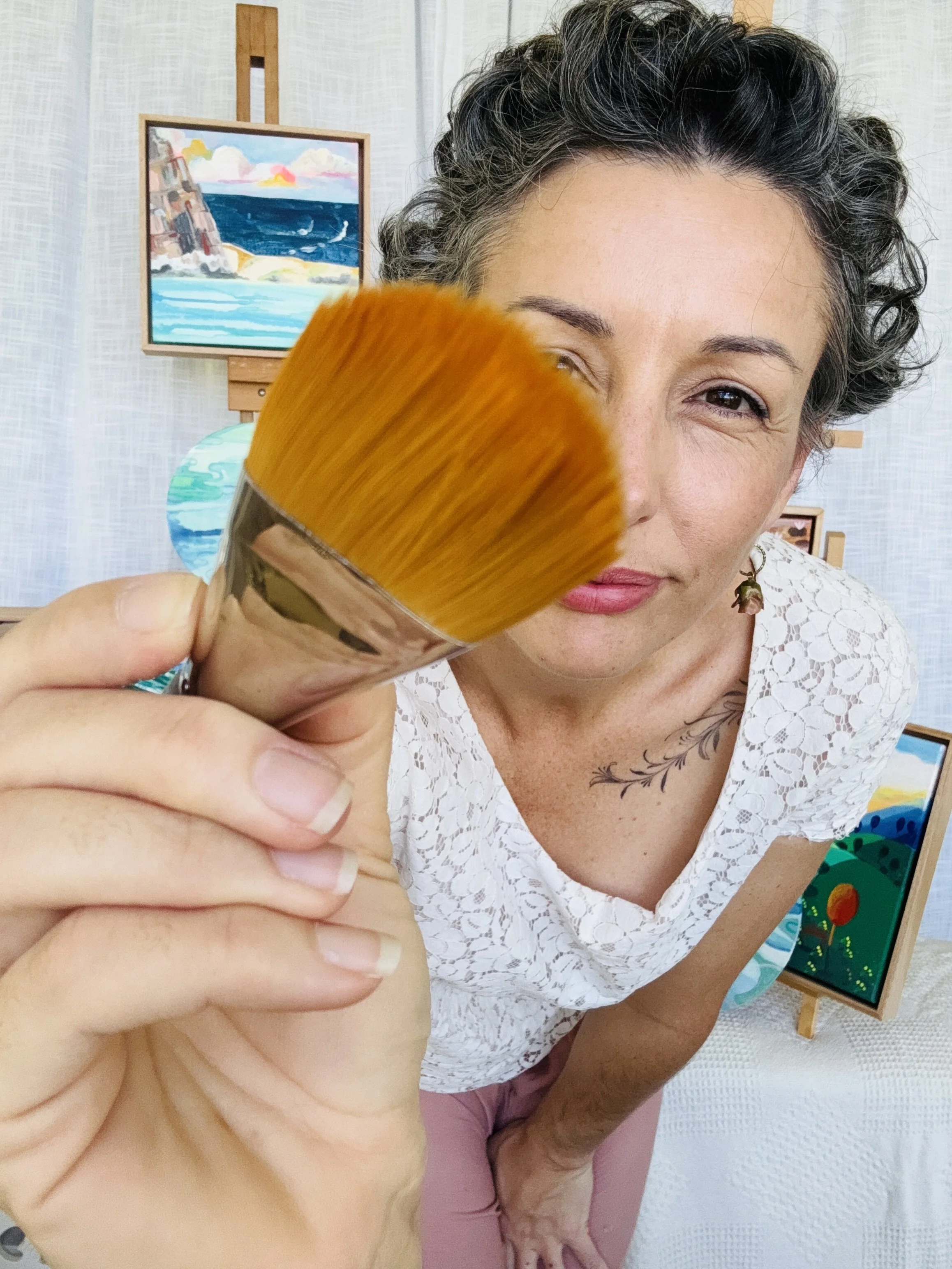 Woman holding a large artists brush close to the camera, with paintings in the background.