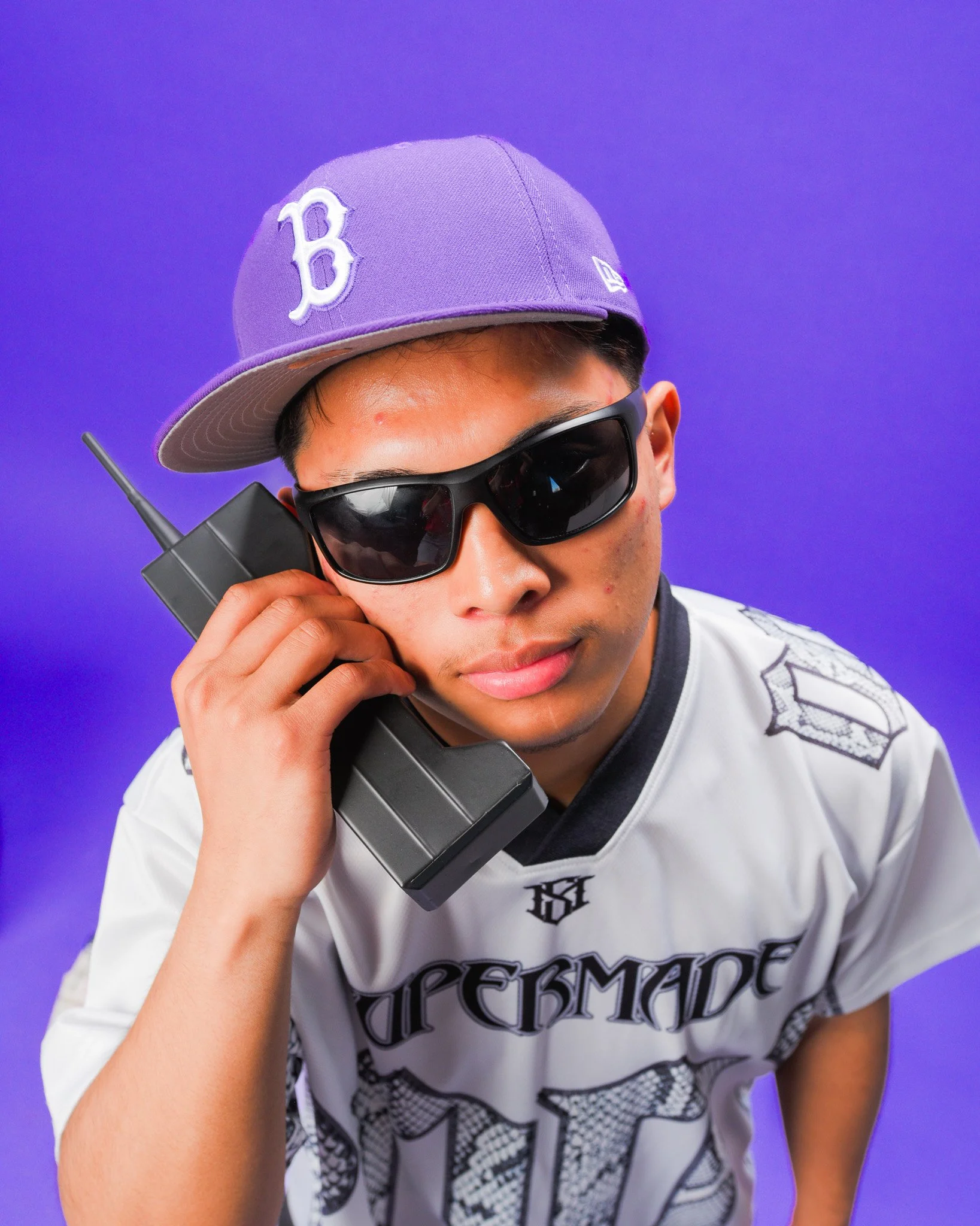 A young man with tan skin wearing black sunglasses and a purple baseball cap with a white logo, holding a walkie-talkie close to his ear against a solid purple background.