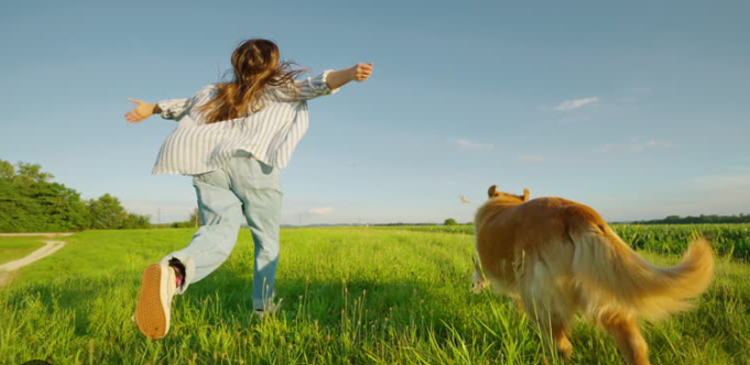 A girl running and playing with a dog in a grassy field under a blue sky.