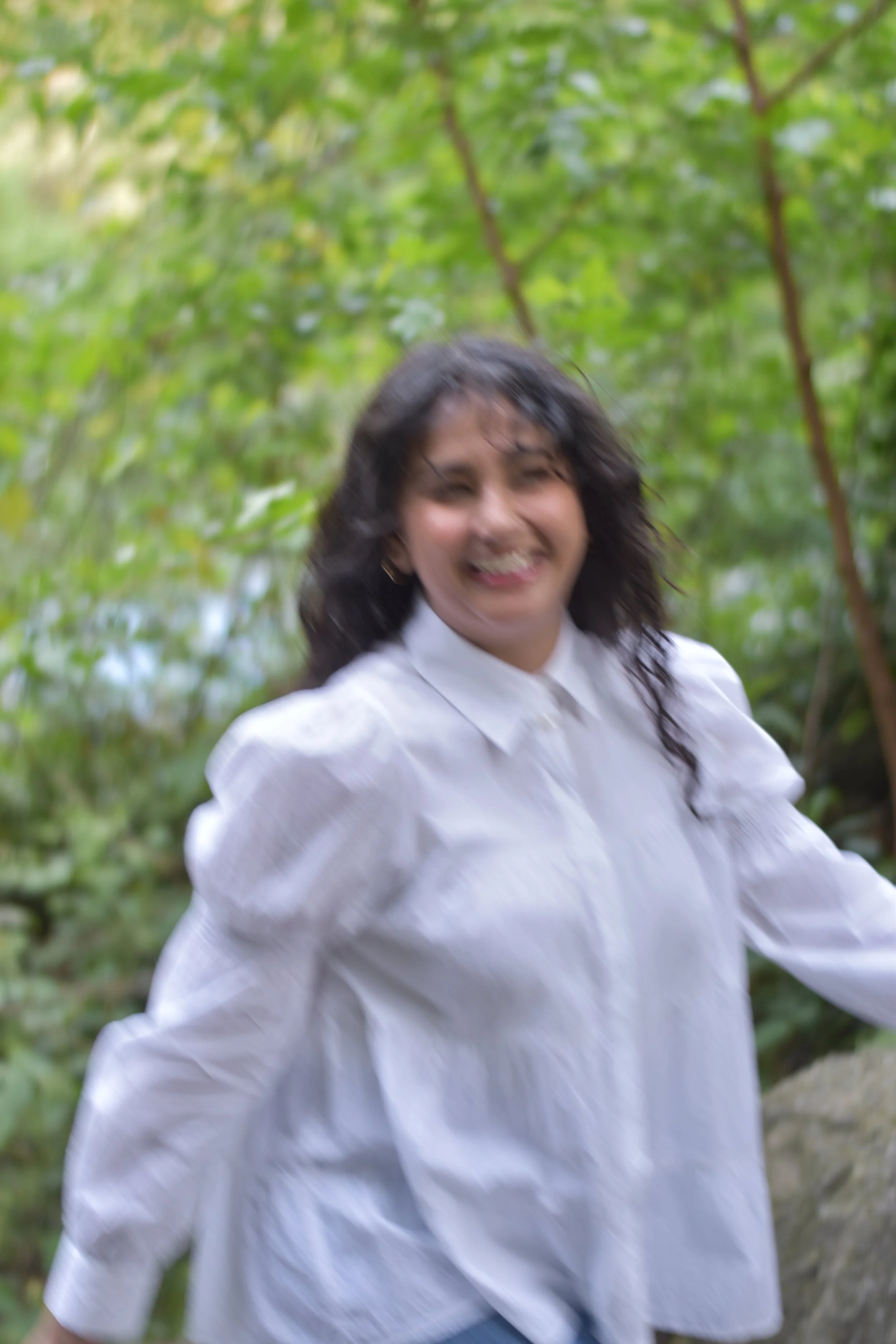 A woman with dark, curly hair smiling and wearing a white shirt outdoors with green trees in the background - head planner Aishee Das