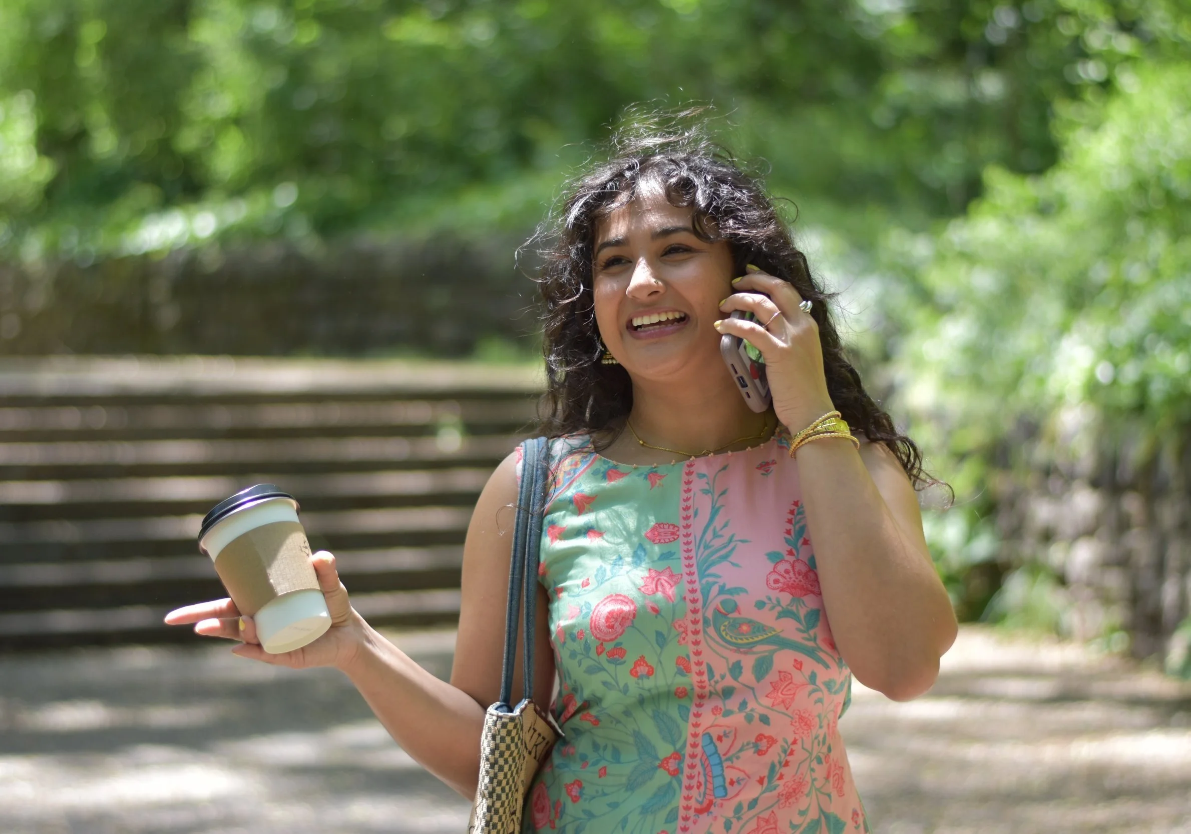 Event and Wedding planner Aishee Das is on the phone laughing as she holds a cup of coffee. She is wearing an Indian print and walking in a park.