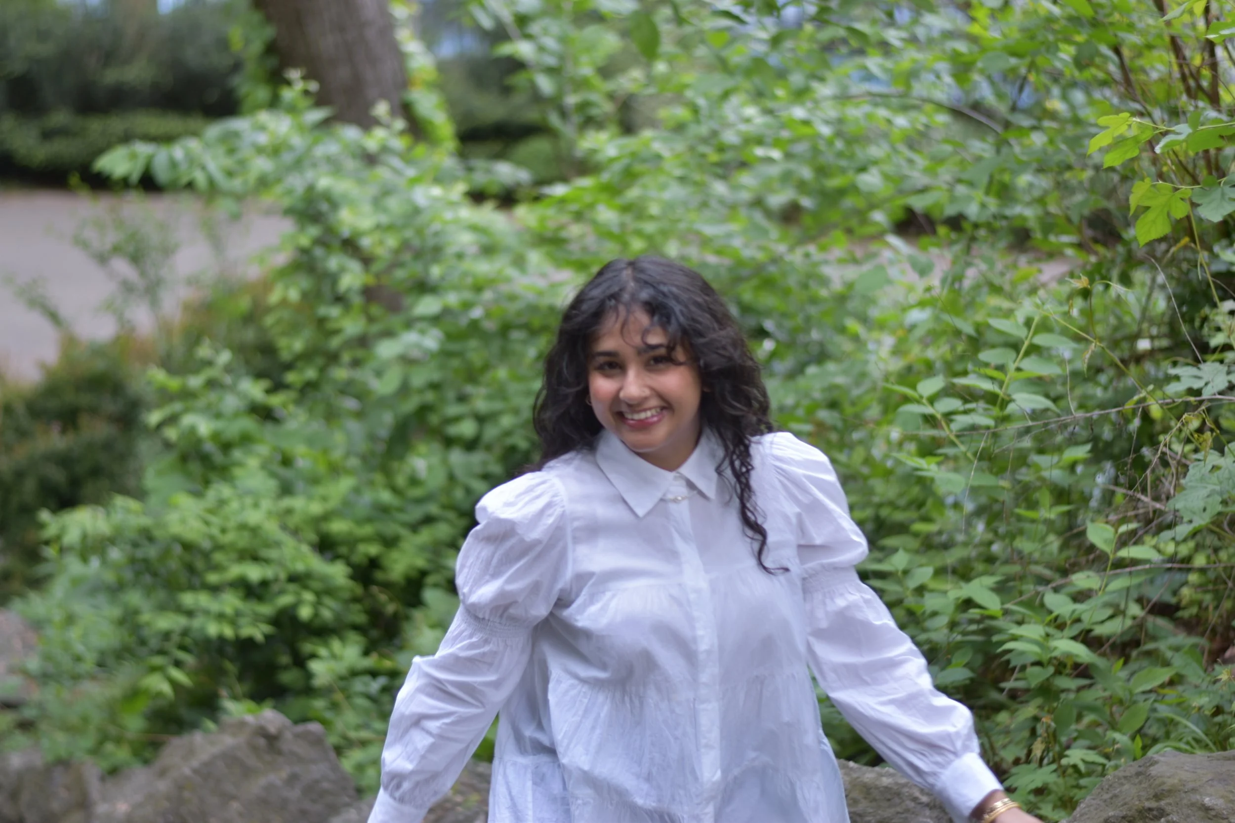 A woman with dark curly hair wearing a white blouse smiling outdoors among green foliage and rocks - head planner Aishee Das