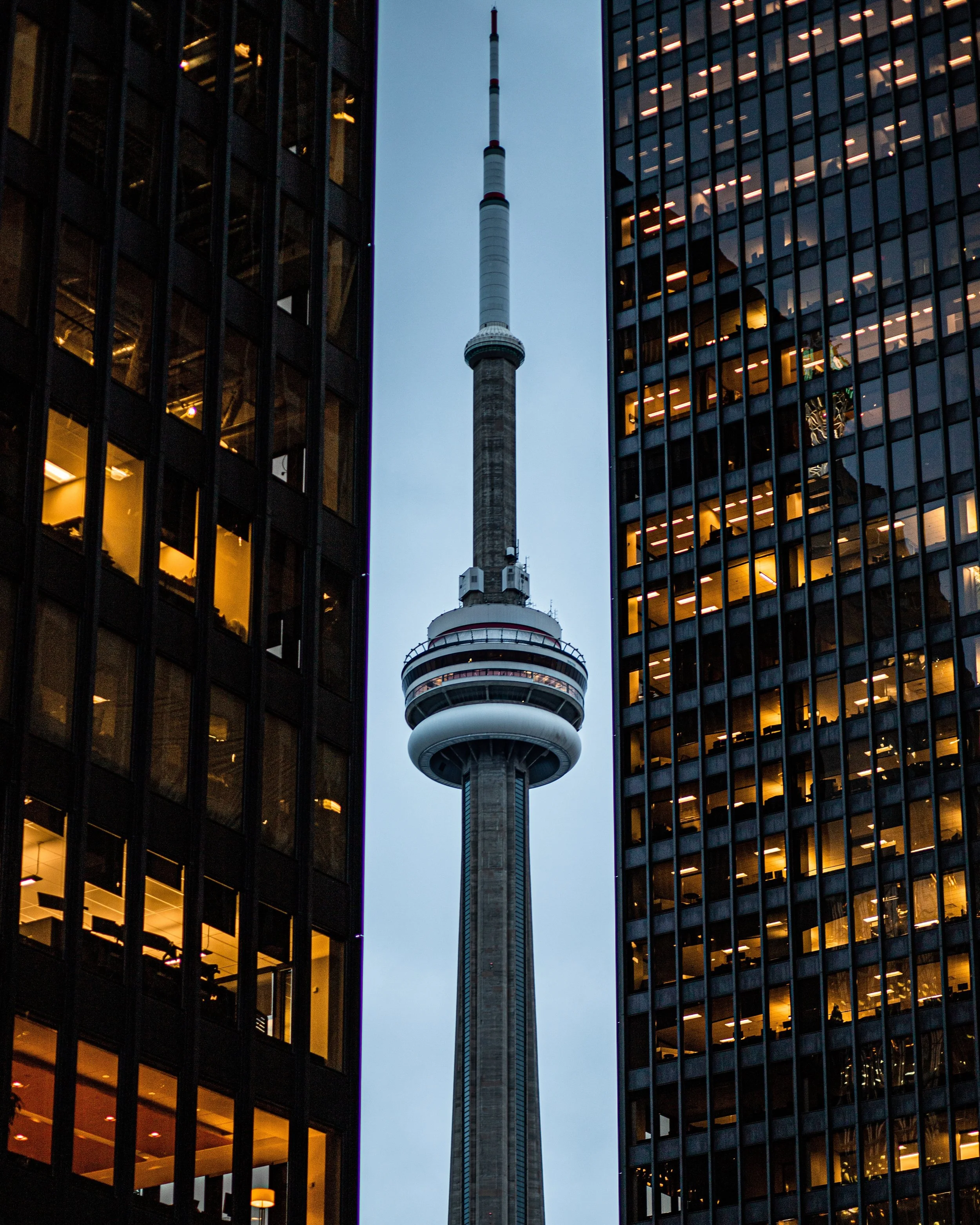CN Tower between two glass office buildings with lit windows at dusk.