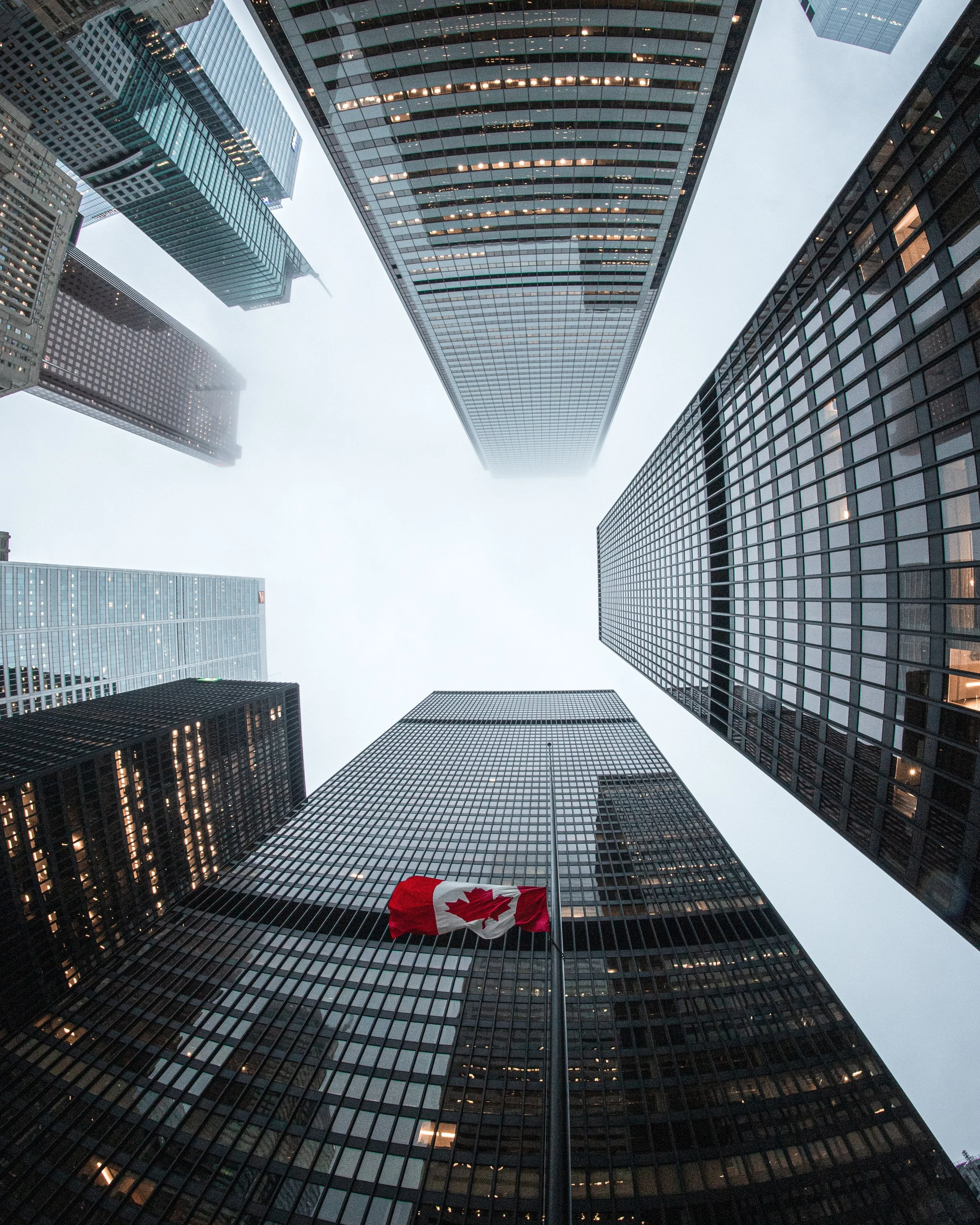 Looking up at tall skyscrapers in a city with a Canadian flag on a flagpole in the foreground.