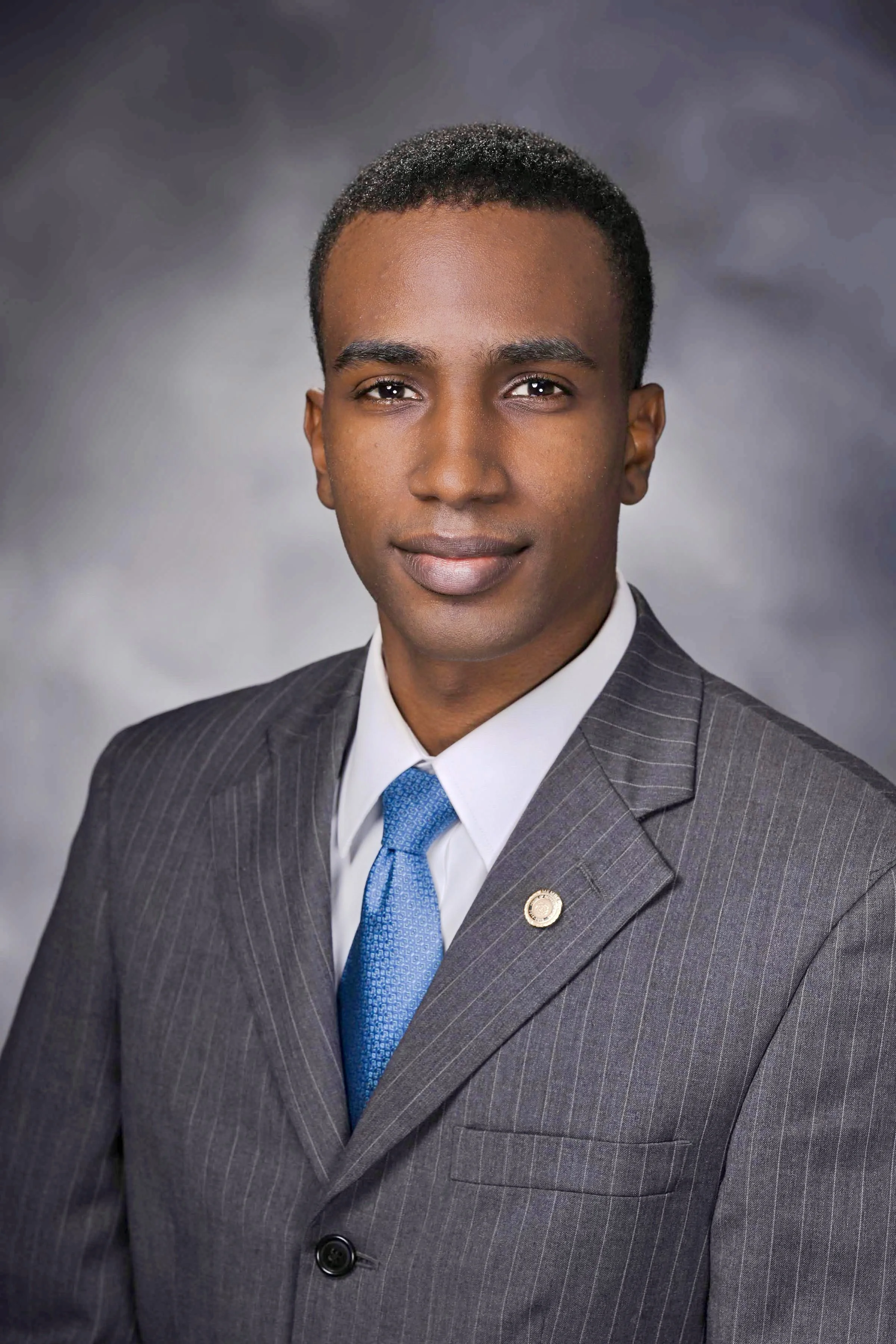 Professional headshot of a man in a gray pinstripe suit, white shirt, and blue tie, against a gray background.