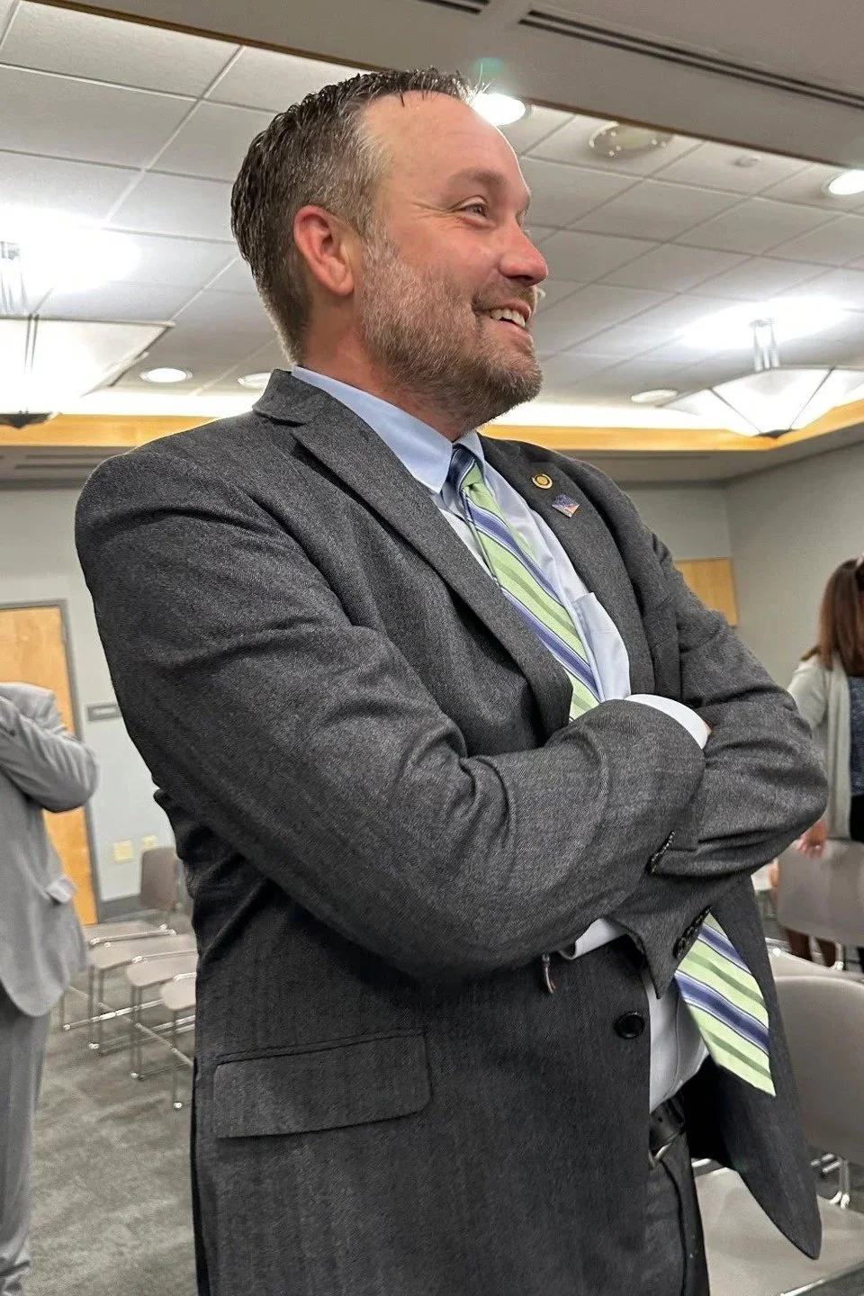 A man in a gray suit with a light green striped tie, standing with his arms crossed, smiling, in a conference room with a few other people in the background.