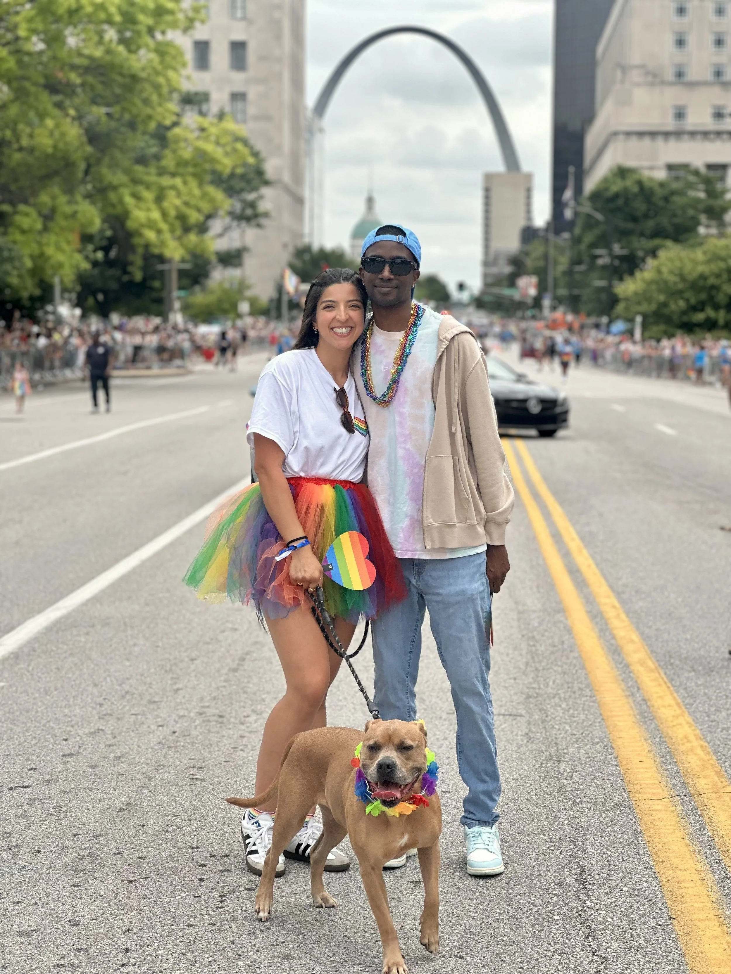 Two people wearing rainbow-themed clothing and accessories stand in the middle of a busy street parade, with a dog wearing a rainbow lei and colorful collar, and the Gateway Arch in the background.