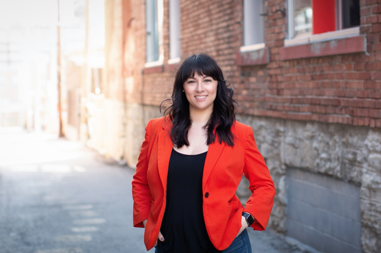 A woman with dark hair and bangs standing outdoors on a sidewalk with brick buildings in the background, wearing a bright orange blazer and black top, smiling at the camera.