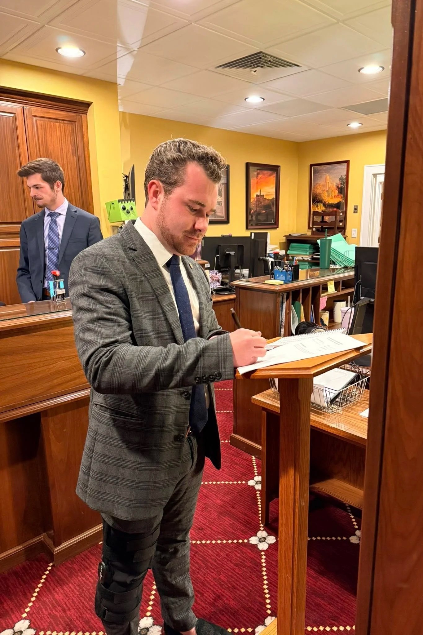 Man in a gray plaid suit signing documents at a desk in a formal office, with another man in a suit standing in the background.