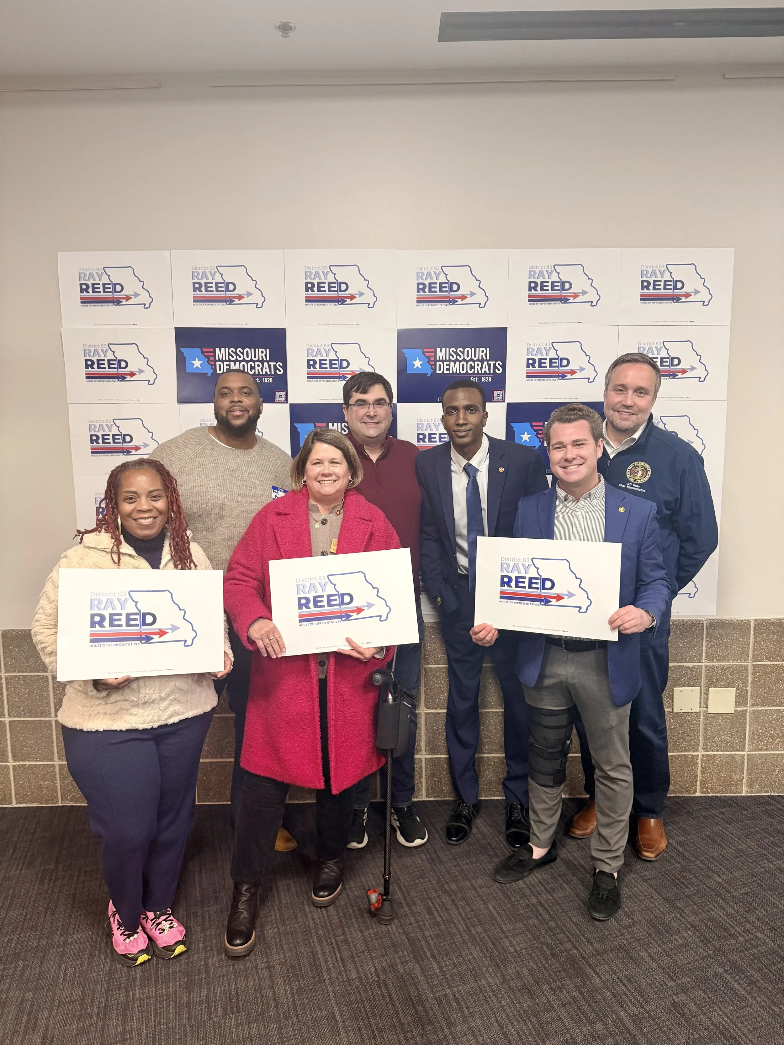 Group of seven diverse people standing in front of a backdrop with Missouri Democrats and Ray Reed logos, holding signs with Ray Reed's name and state outline.