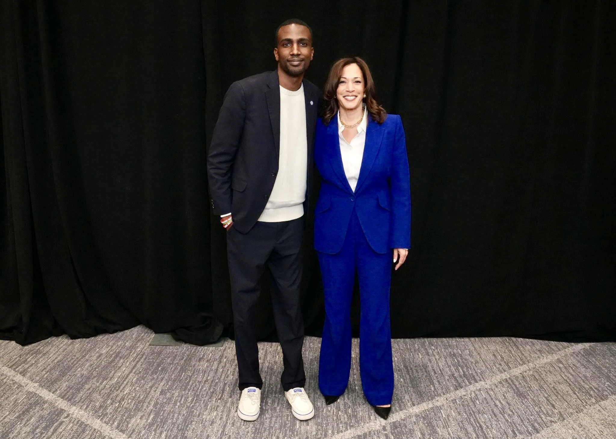 A man and a woman standing side by side, smiling in front of a black curtain backdrop. The man is wearing a dark blazer, white sweatshirt, dark pants, and white sneakers. The woman is dressed in a bright blue pantsuit with a white blouse and black high heels.