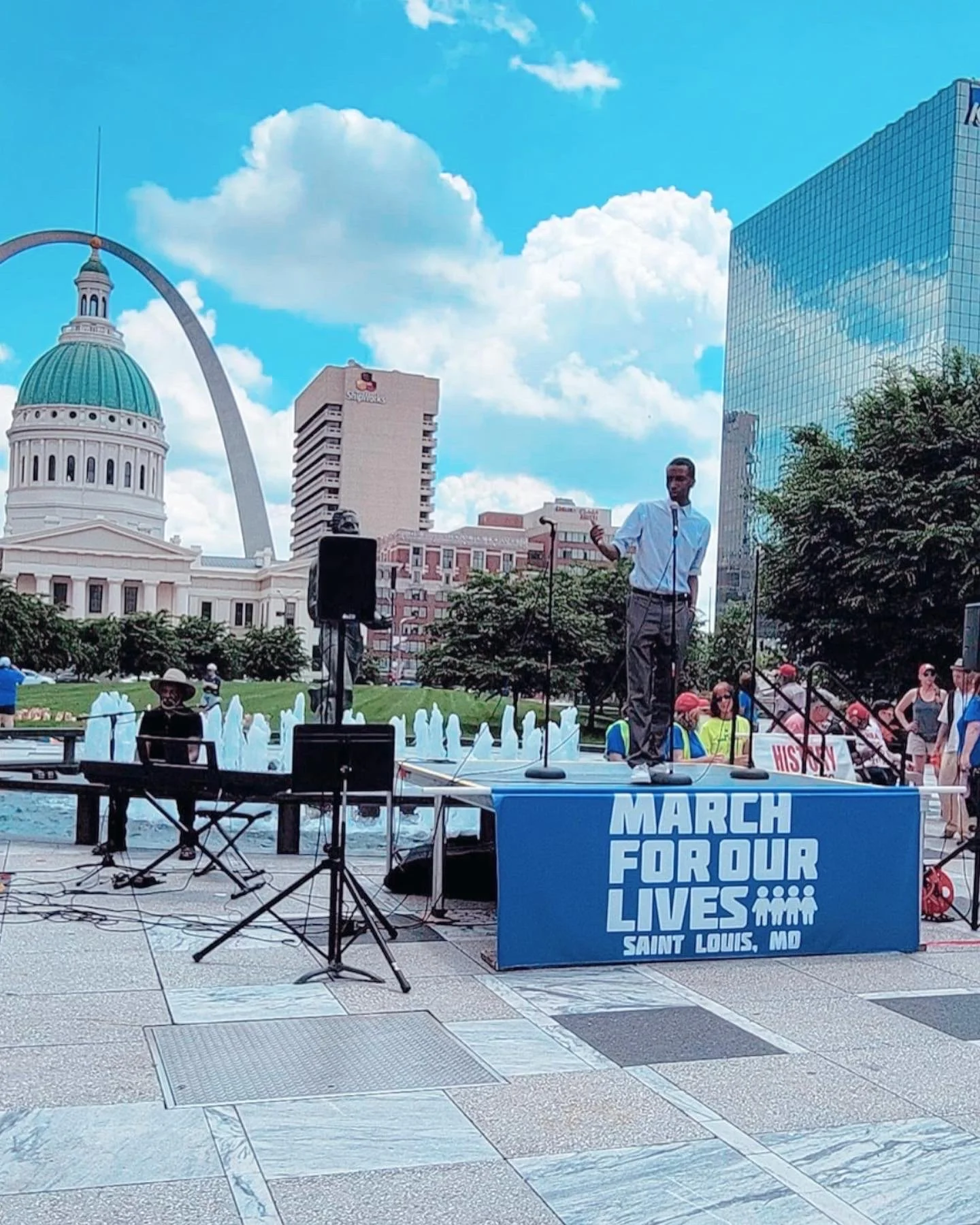A man is speaking into a microphone on a stage with a banner reading 'March for Our Lives, Saint Louis, MO'. There are people gathered around, some with red hats, and a person playing keyboard to the left. The background features the Gateway Arch, a historic building with a dome, and modern glass buildings under a partly cloudy sky.