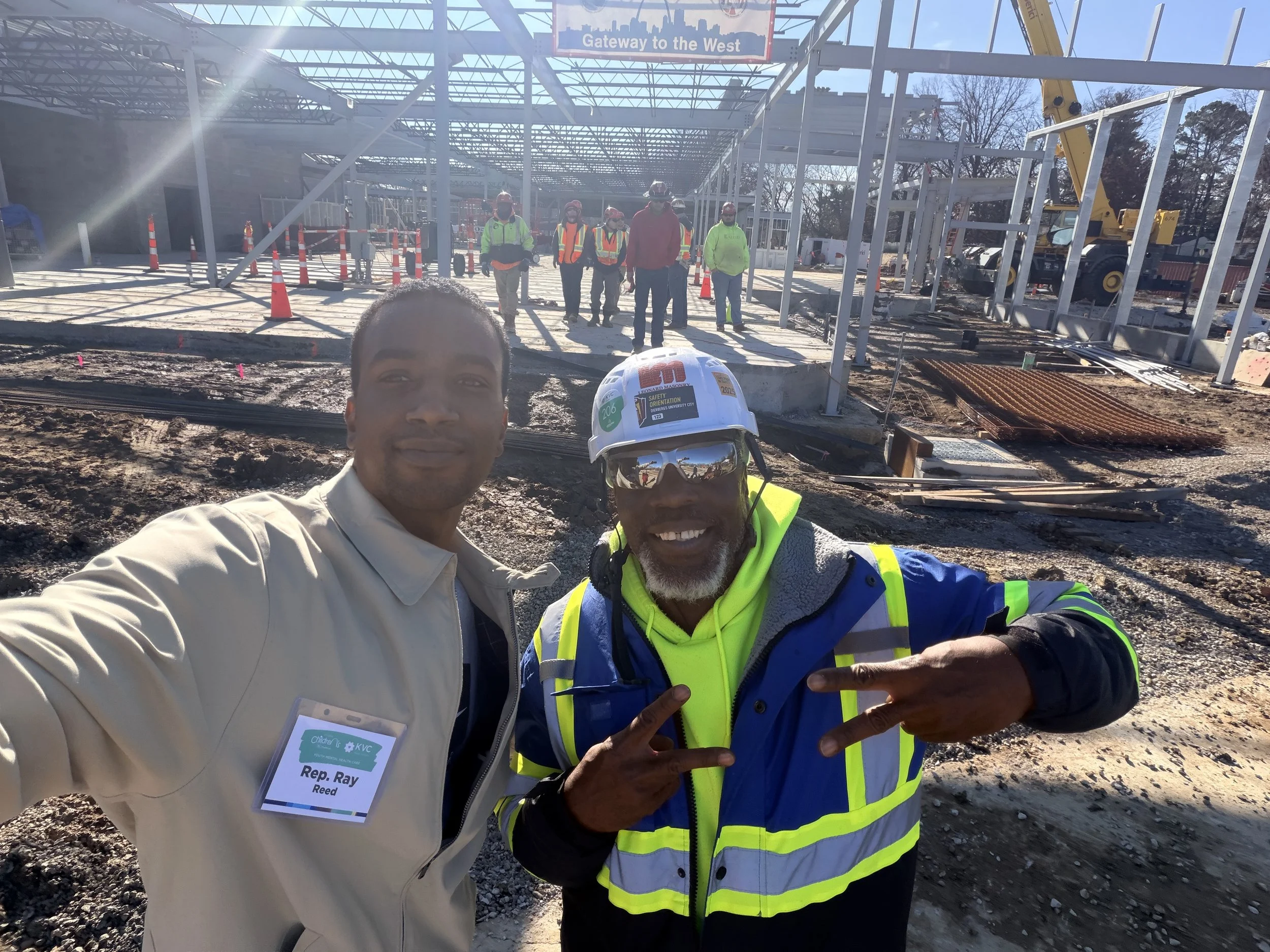 Two men posing for a selfie at a construction site with a partially built structure in the background and several workers in safety gear. One man is wearing a light-colored jacket with a name tag, and the other is wearing a safety helmet and jacket, making peace signs.