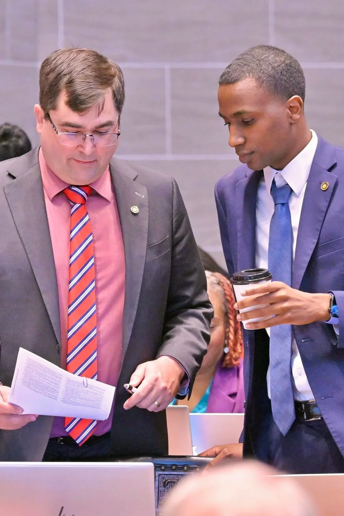 Two men in business suits are looking at a document and a laptop on a table during a meeting or conference.