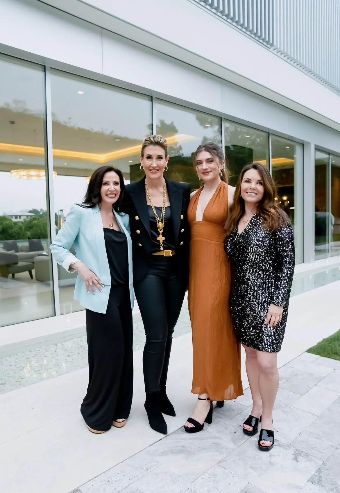 Four women standing outside a modern building, dressed in stylish outfits, smiling for the photo.