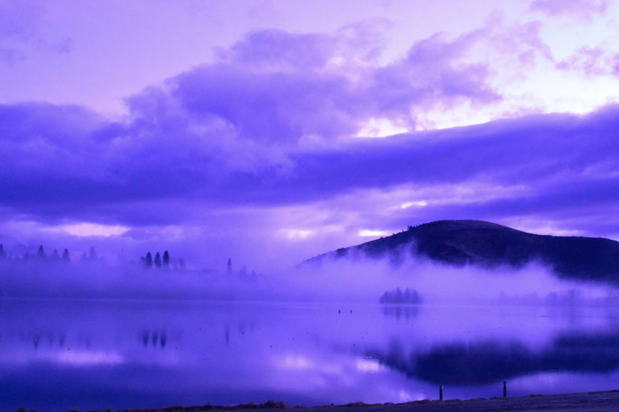 Serene lake scene at dawn with purple and blue sky, mist over water, distant trees, and hills in the background.