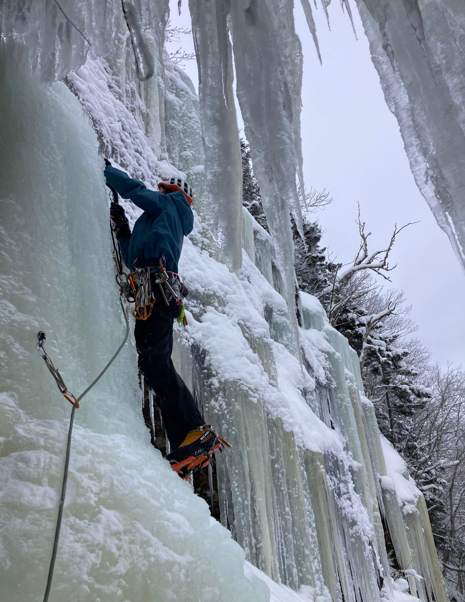 A person ice climbing on a frozen waterfall with ice tools and crampons, wearing a helmet and harness in a snowy, icy landscape.
