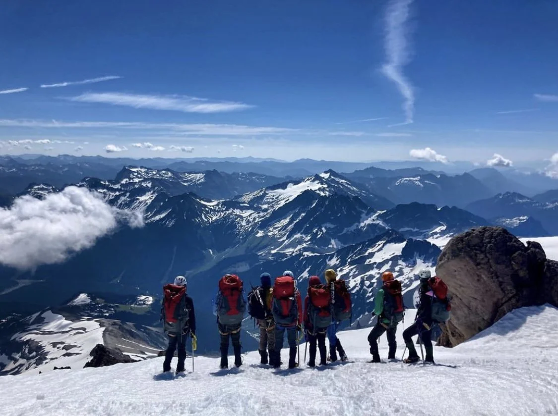 Group of climbers with backpacks and gear standing on snow-covered mountain, overlooking snow-capped peaks and valleys under a partly cloudy sky.