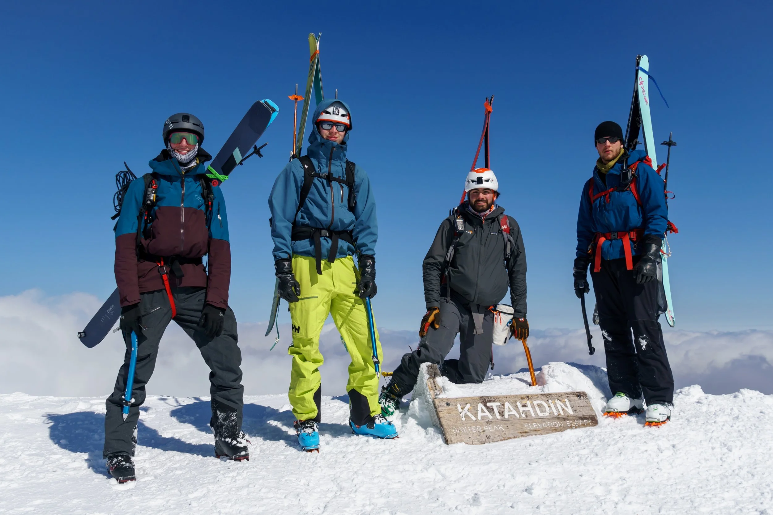 Four mountaineers standing on snow-covered peak with mountain gear. Two are holding skis, one has an ice axe, and one has snowboarding gear. Behind them is a wooden sign that reads "KATAHDIN, Baxter Peak, Elevation 5,267 ft." against a backdrop of blue sky and clouds.