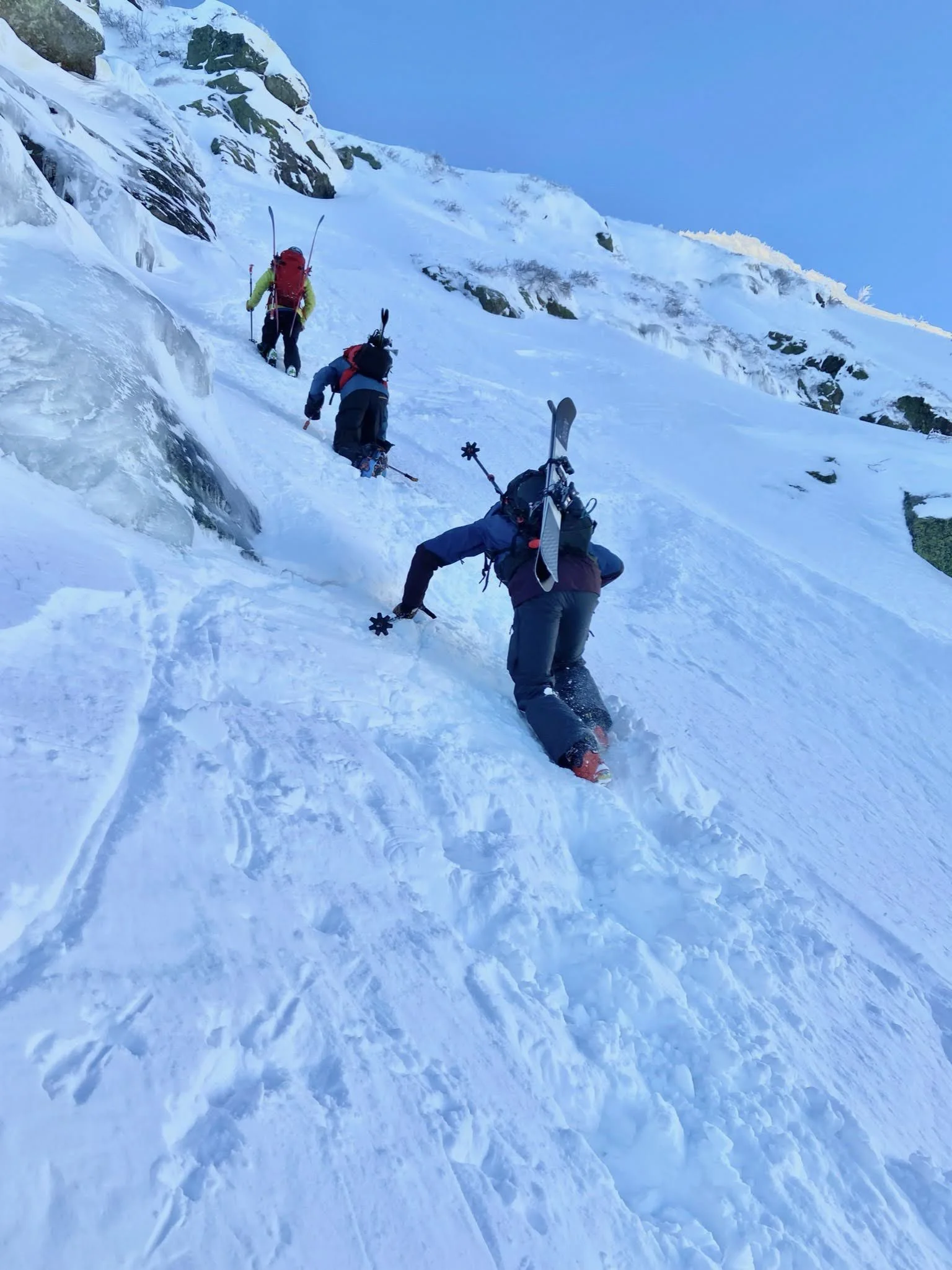 Four people in winter gear climbing a snowy, icy mountain slope with rocky outcrops visible, carrying skis and trekking poles.