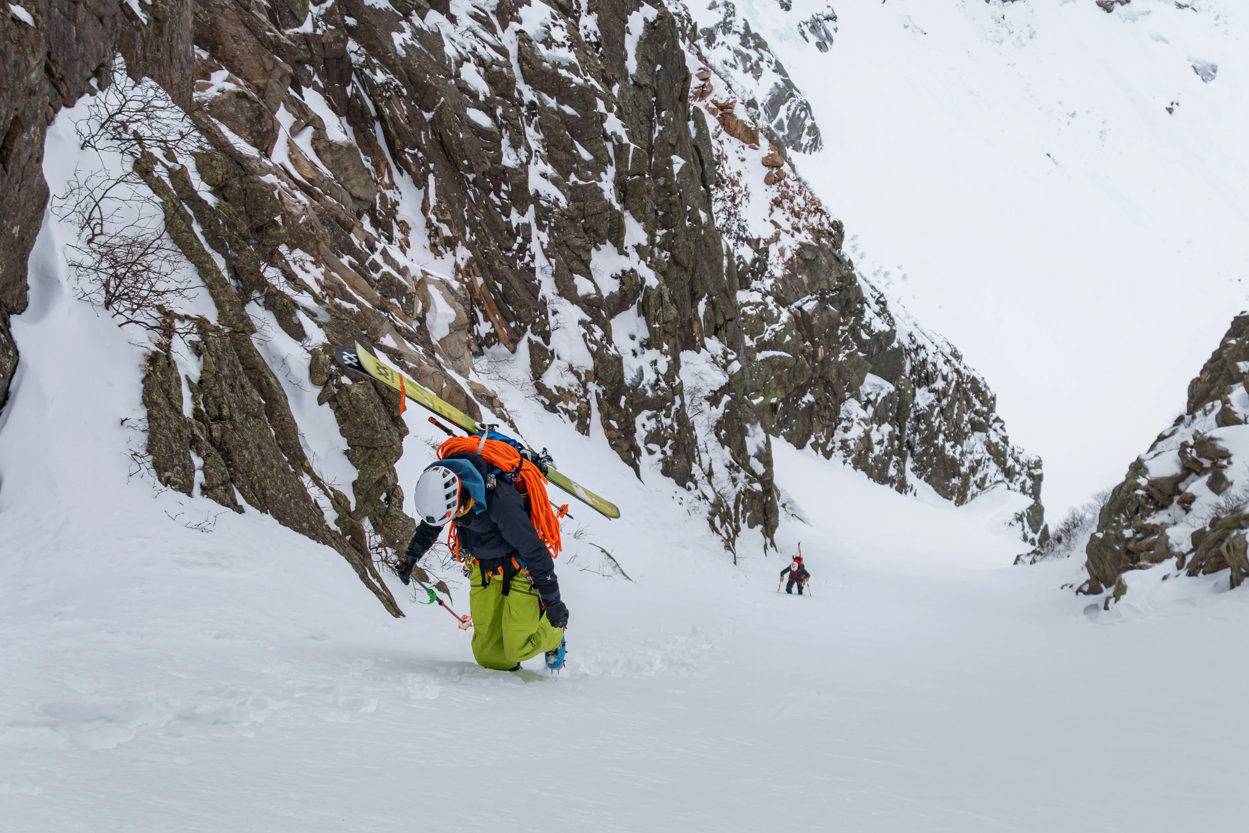Two climbers traversing a snowy mountain pass with rocky cliffs and snow-covered slopes.