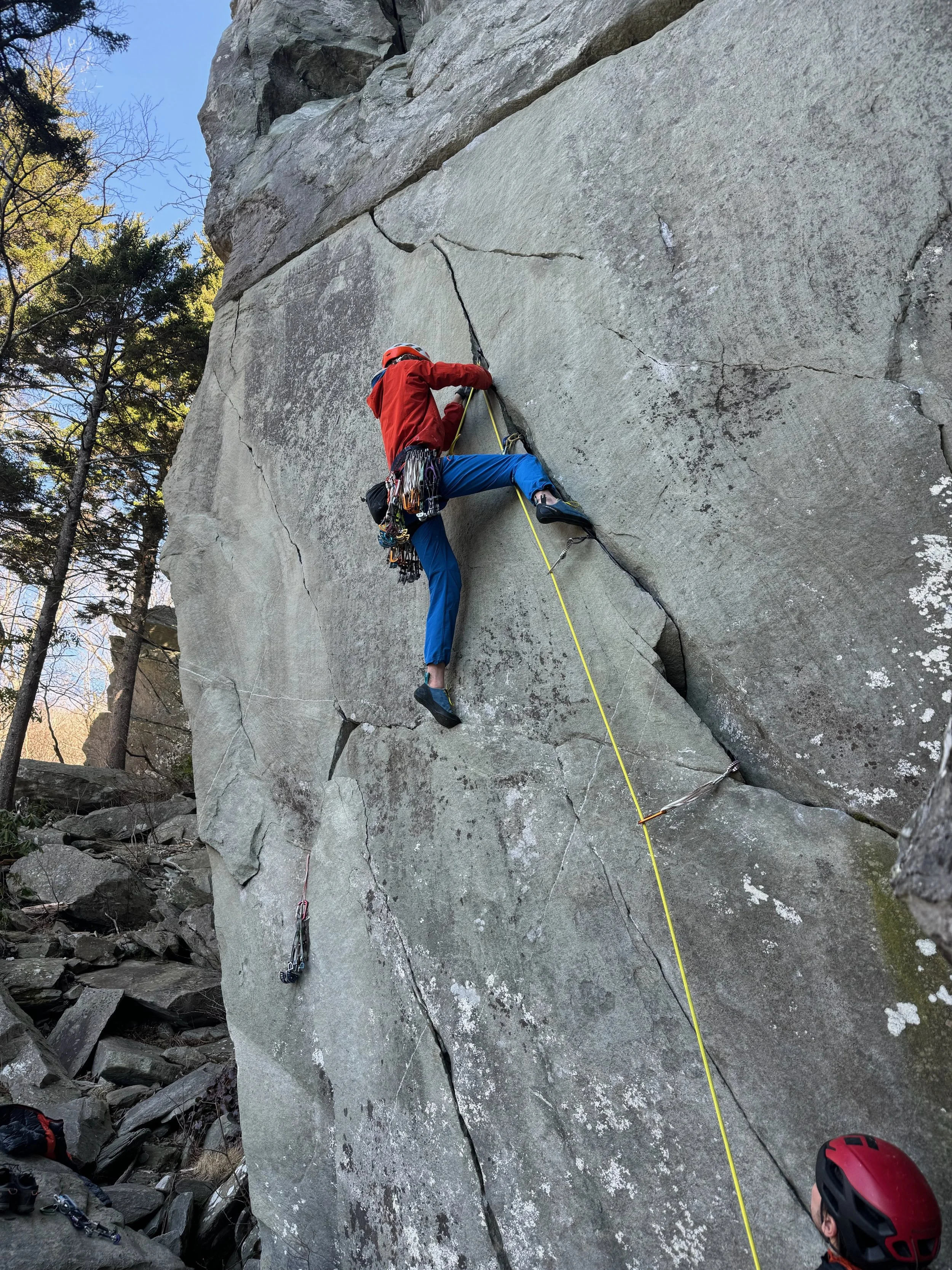A person rock climbing on a large outdoor granite wall with trees and rocks nearby, wearing a red jacket, blue pants, and a helmet, secured with safety ropes.